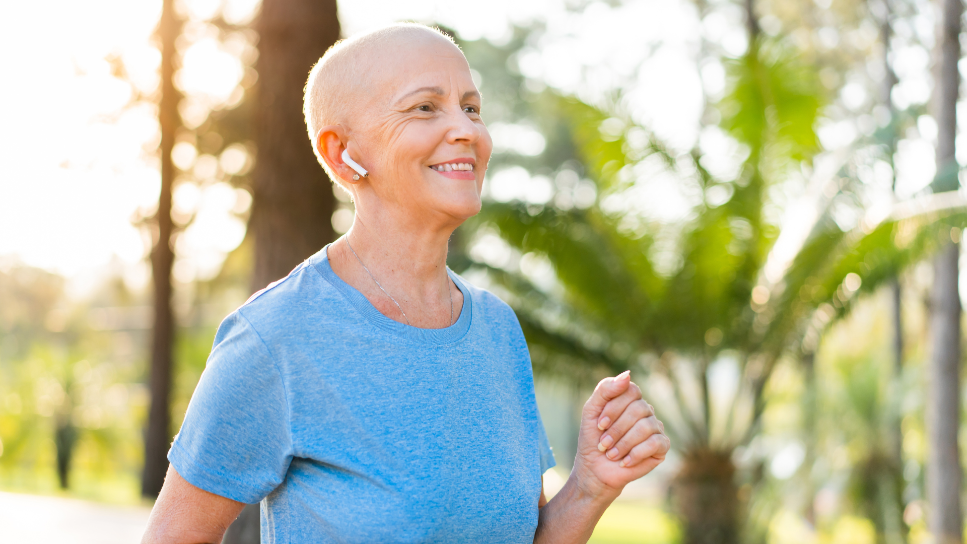 Woman with a shaved head smiles while jogging outdoors; wearing earbuds and a blue shirt.