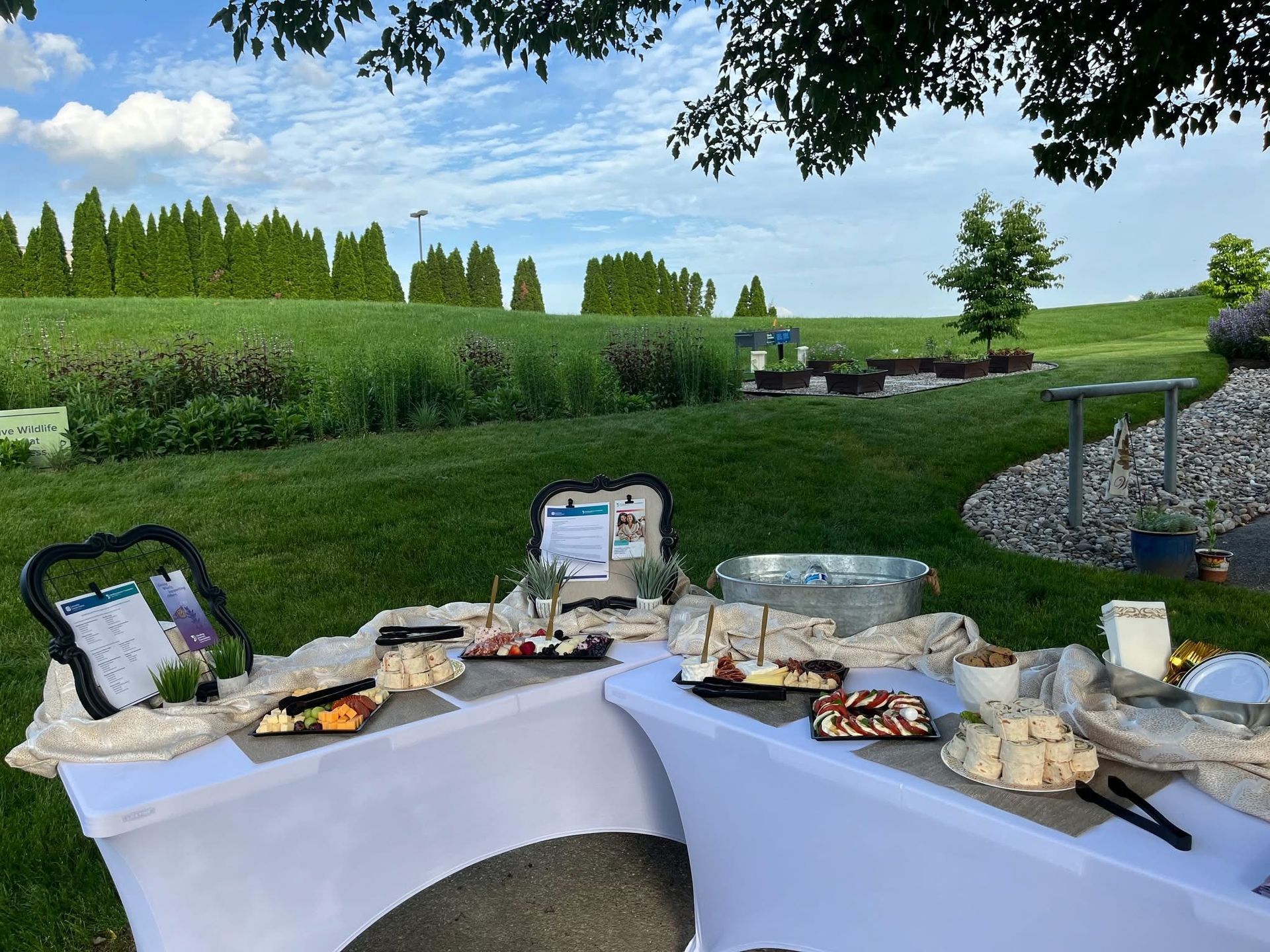 Two white-clothed tables with food offerings set up outdoors on a lawn with trees and a blue sky.