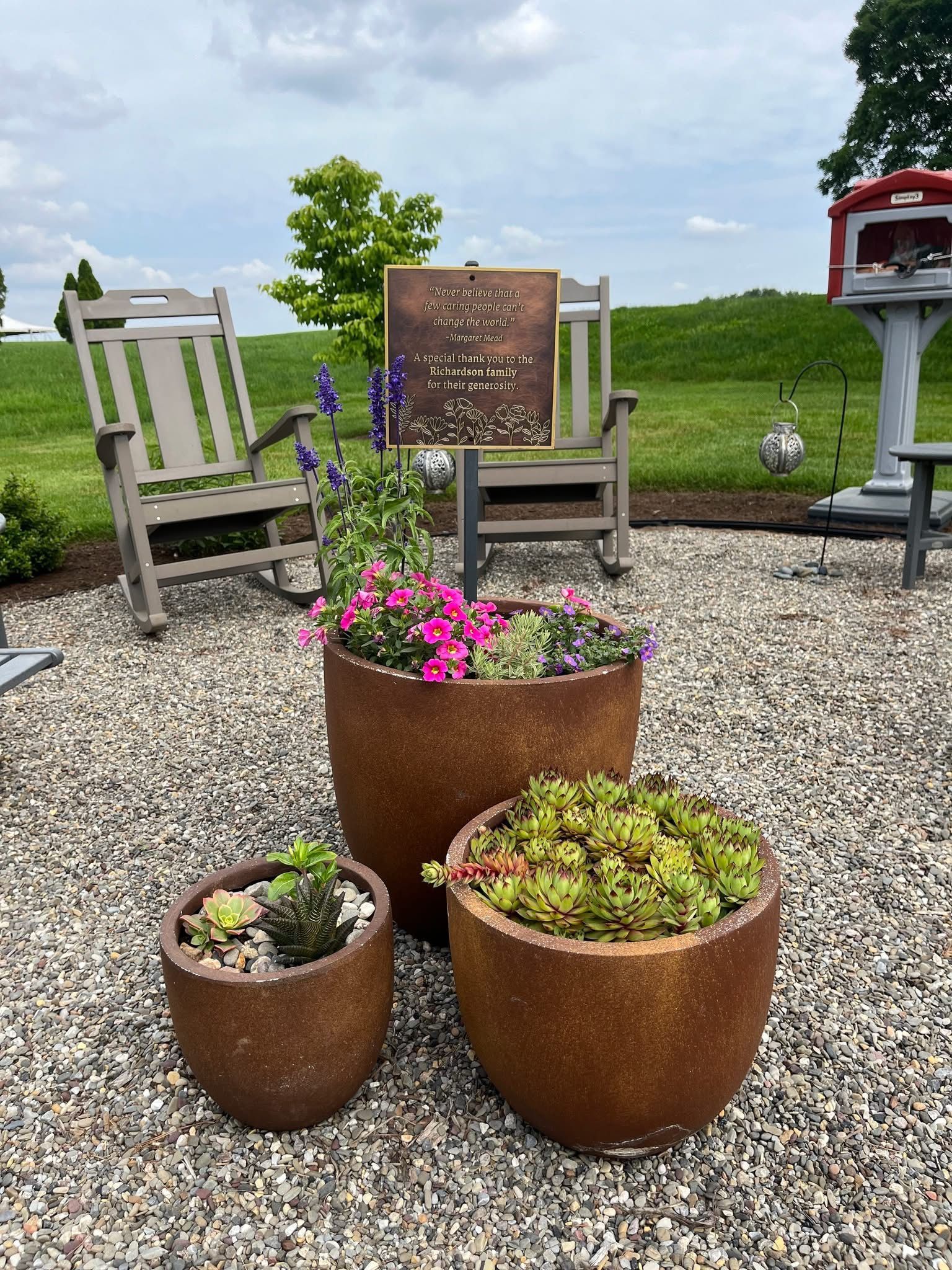 Three brown planters with colorful flowers on a stone patio, two rocking chairs, and a memorial plaque.