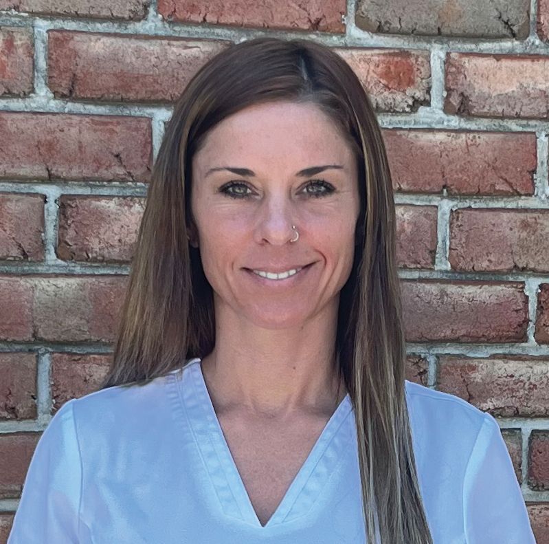 Healing Journey Foundation Board | Woman with long brown hair, wearing a white top, smiling in front of a brick wall.