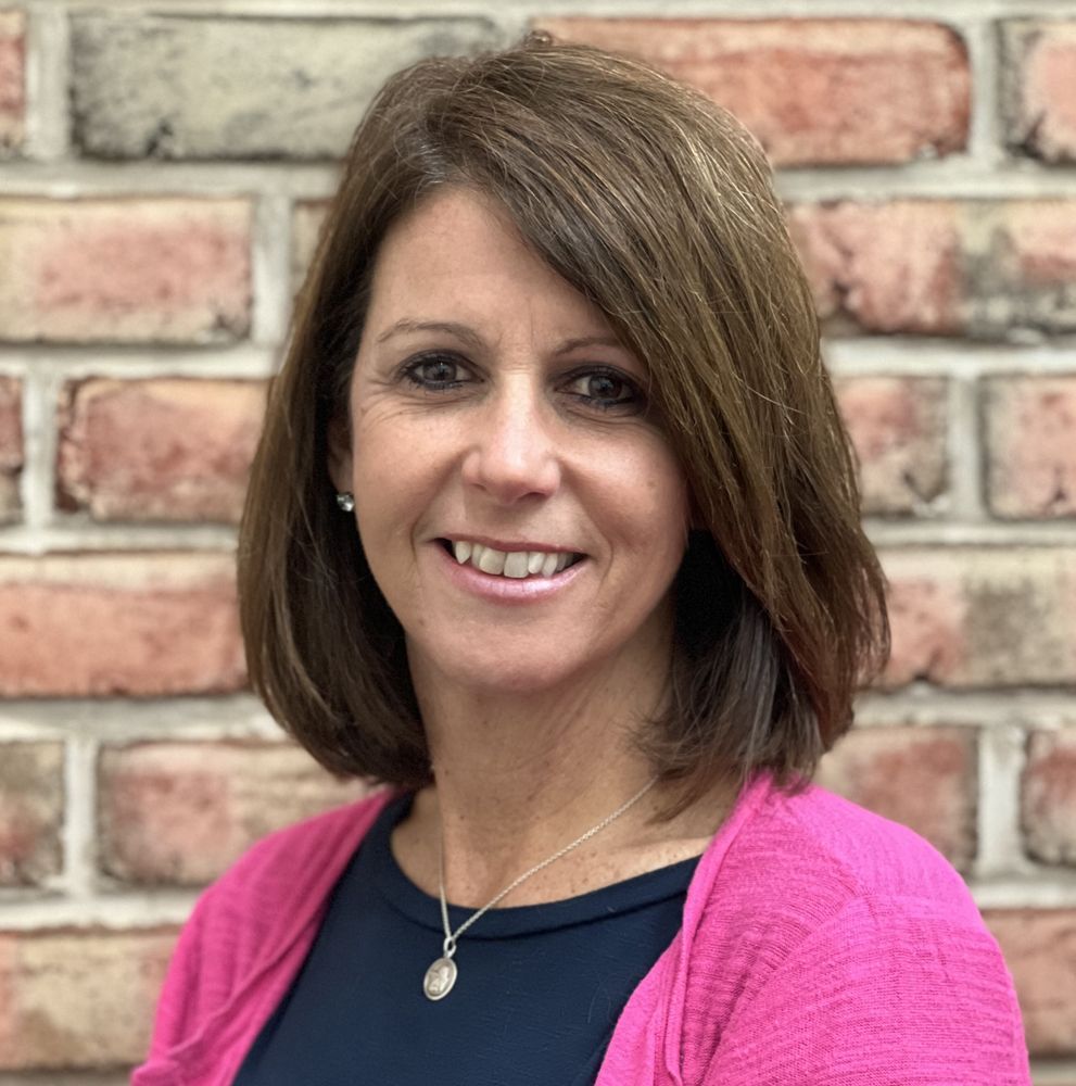 Woman with brown hair and a pink cardigan smiling in front of a brick wall.