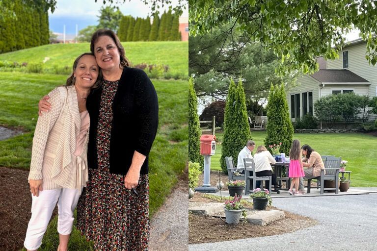 Two women embrace outdoors. A group of people sits around a table. Green lawn, trees, house.