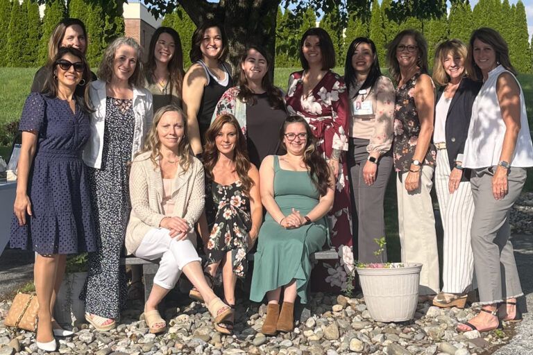 Group of women posing outdoors; some standing, some seated. They are smiling in front of foliage and a building.
