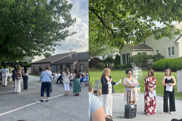 Event outdoors with people listening to speakers. Buildings and trees in background.