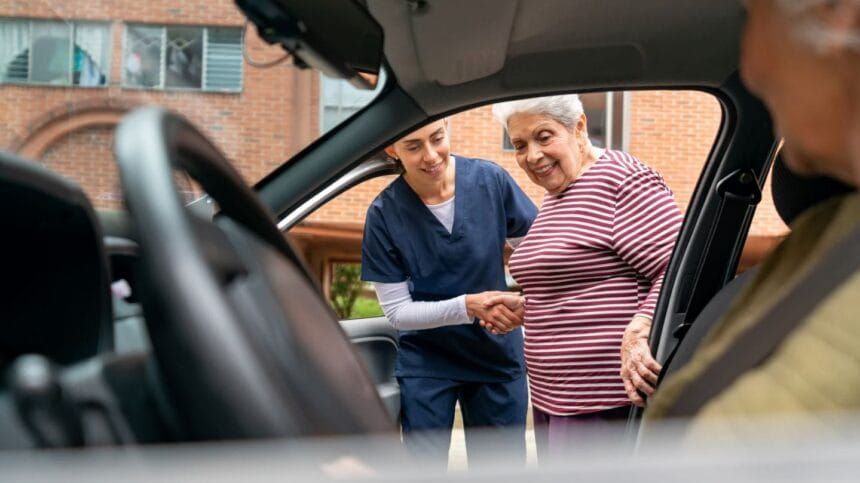 Woman assisting another woman getting into a car; outside a brick building.