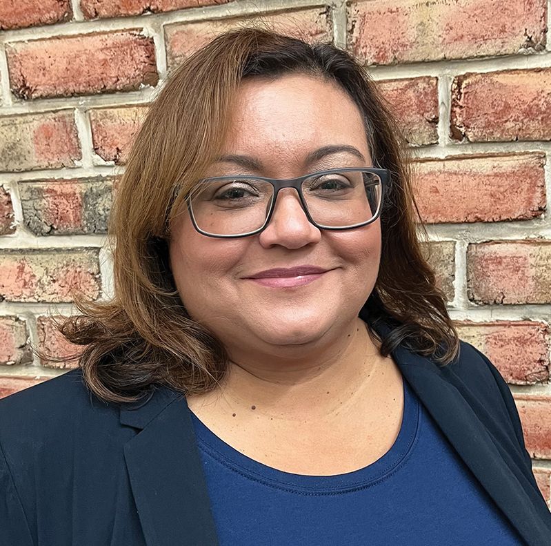 Woman with glasses smiles in front of a brick wall, wearing a navy blazer and blue shirt.