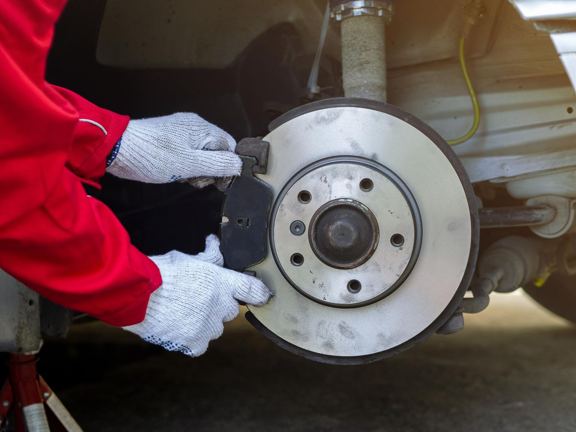 Mechanic in red jacket wearing white gloves removes a car brake rotor near the wheel hub.
