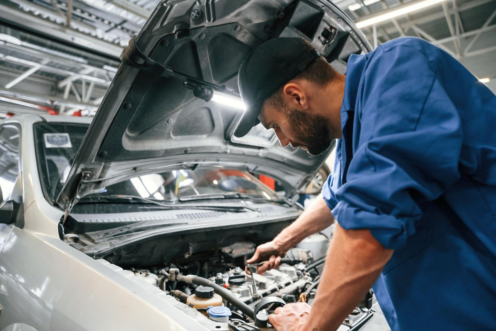 Mechanic in blue coveralls inspecting an engine under an open car hood in a workshop