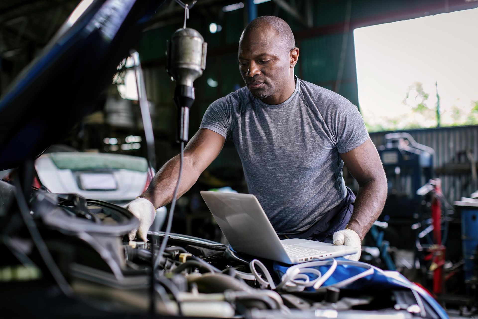 Mechanic working on a car engine in a garage, using a laptop with tools nearby
