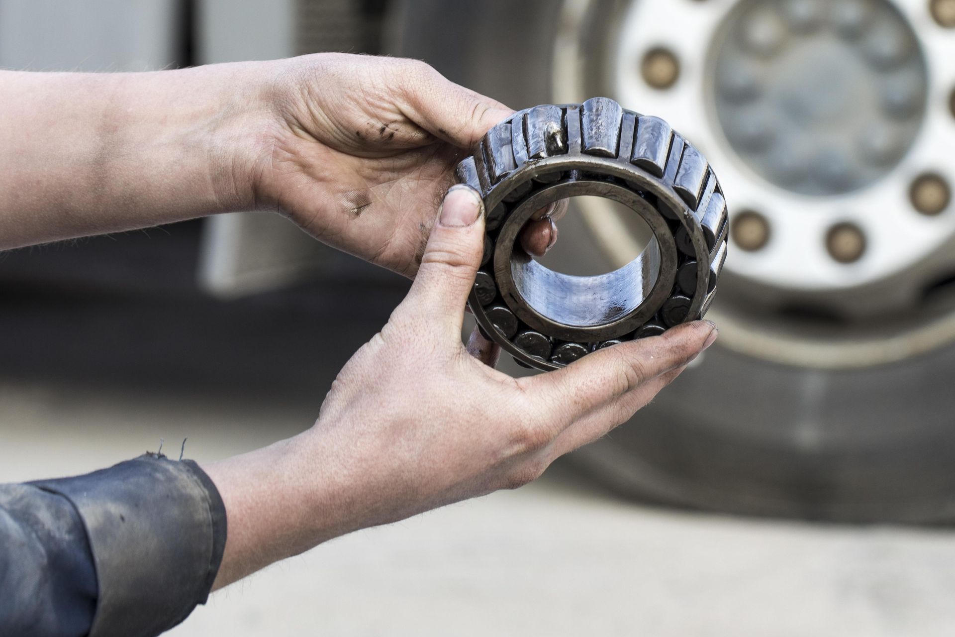 Hands holding a metal bearing or gear part in a workshop, with a car wheel blurred in the background