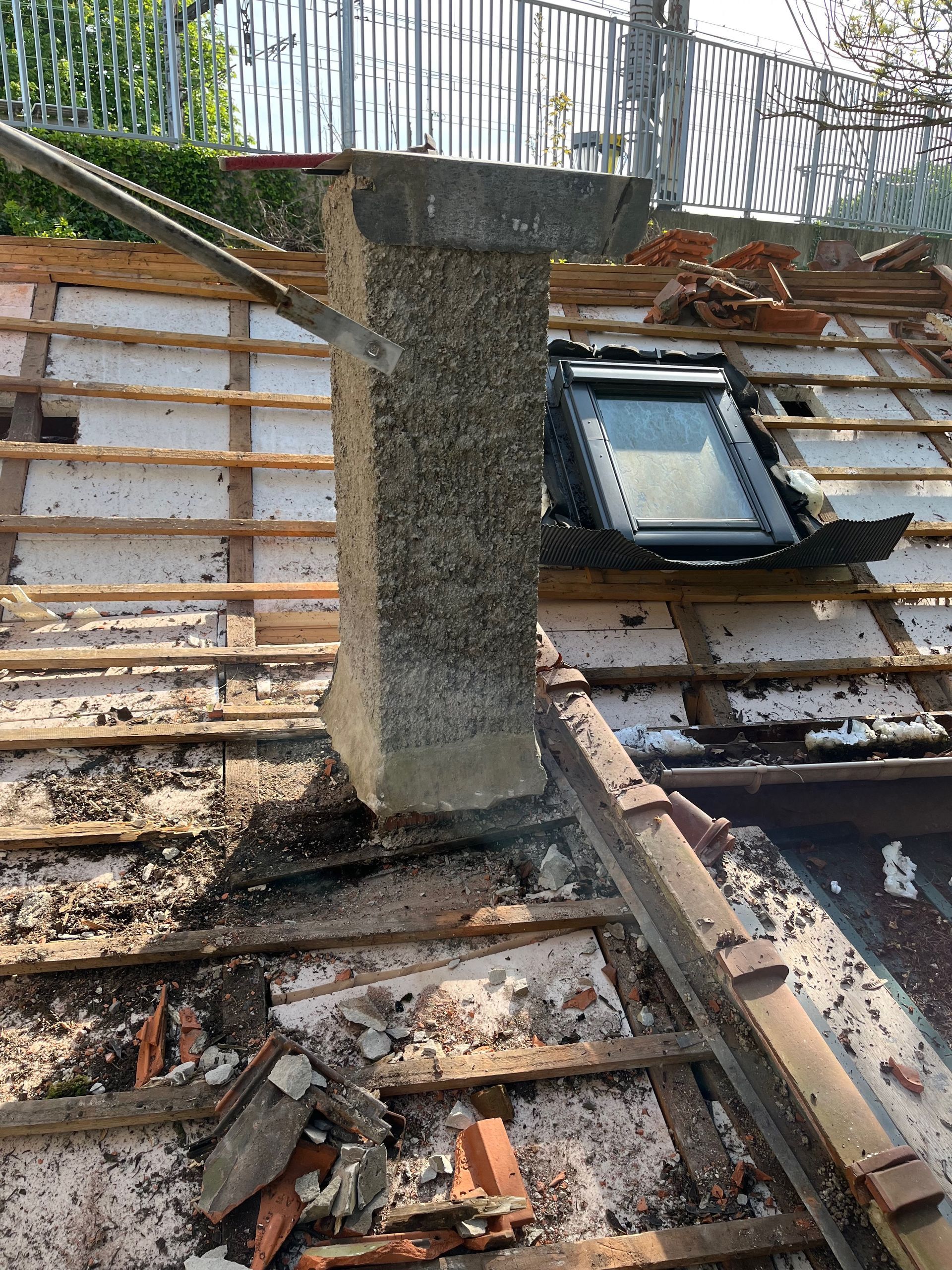 Cheminée sur un toit en construction ; bardeaux endommagés, chevrons apparents, puits de lumière.
