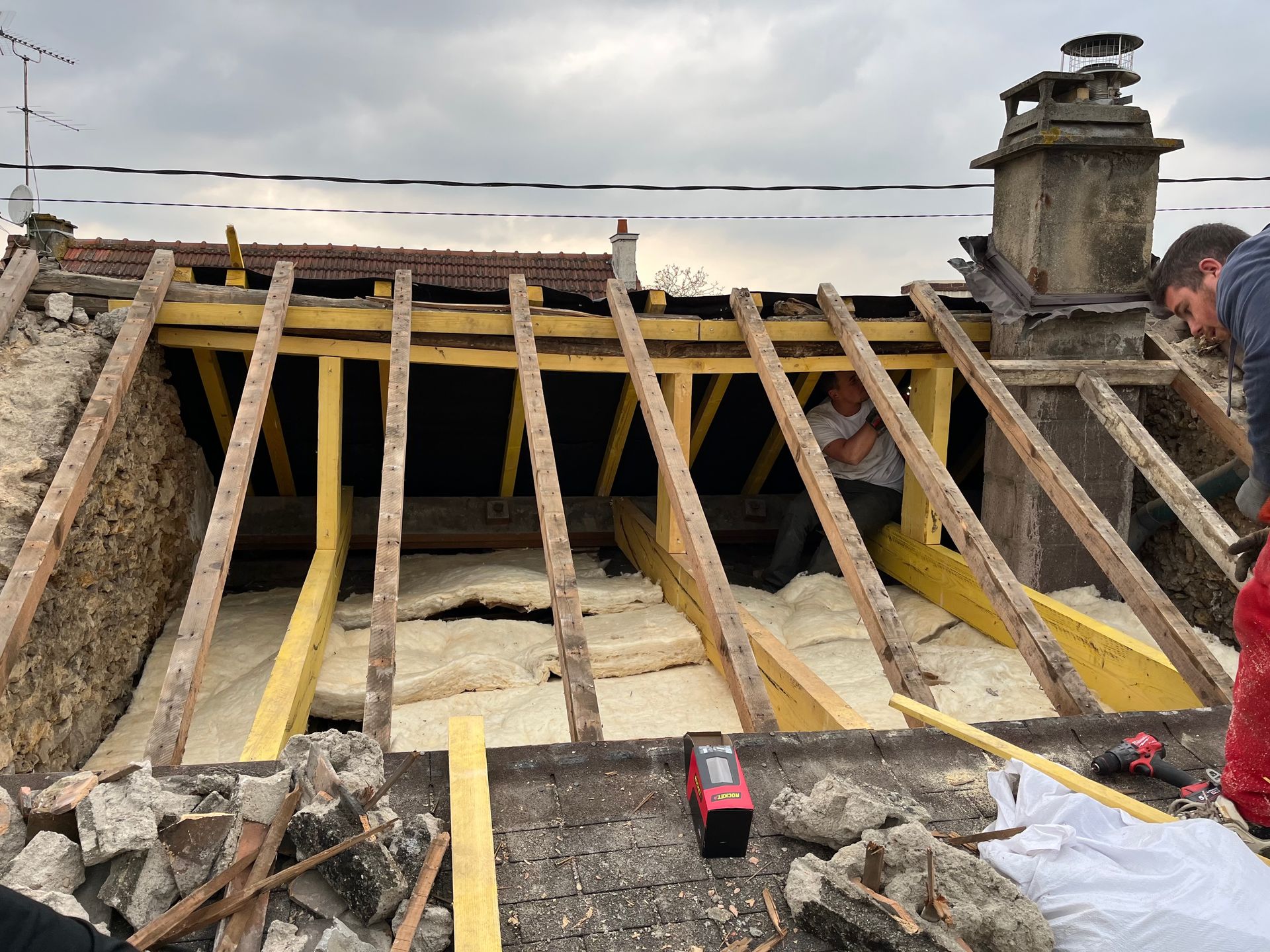 Travaux de toiture : Des ouvriers installent de l'isolant entre les chevrons d'un toit endommagé, avec une cheminée et un ciel nuageux.