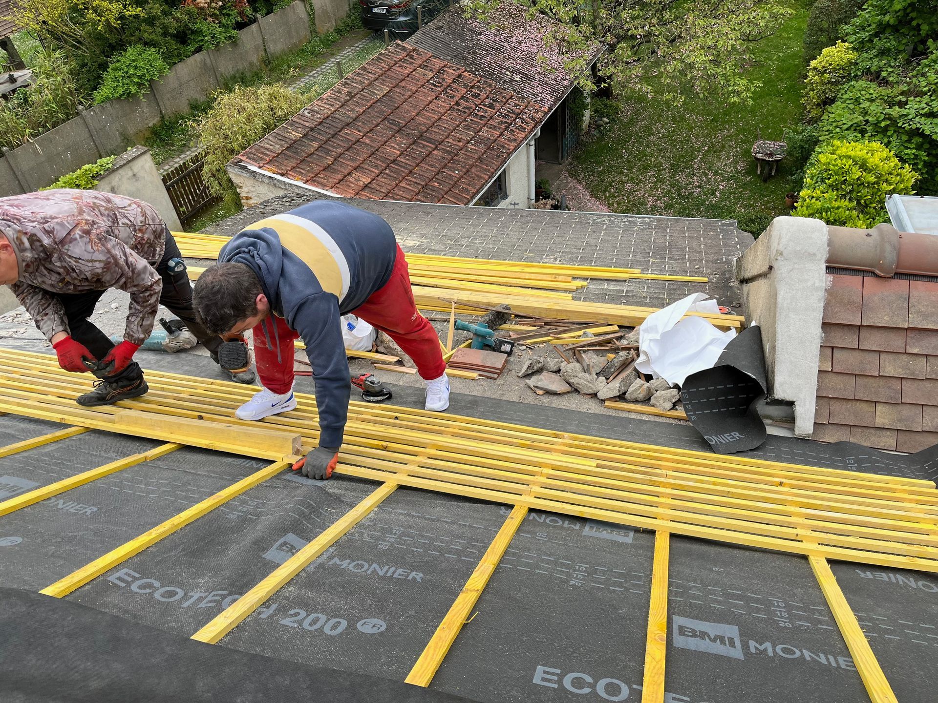 Deux personnes installent des linteau jaune sur un toit noir. Des outils et une cheminée se trouvent à proximité.