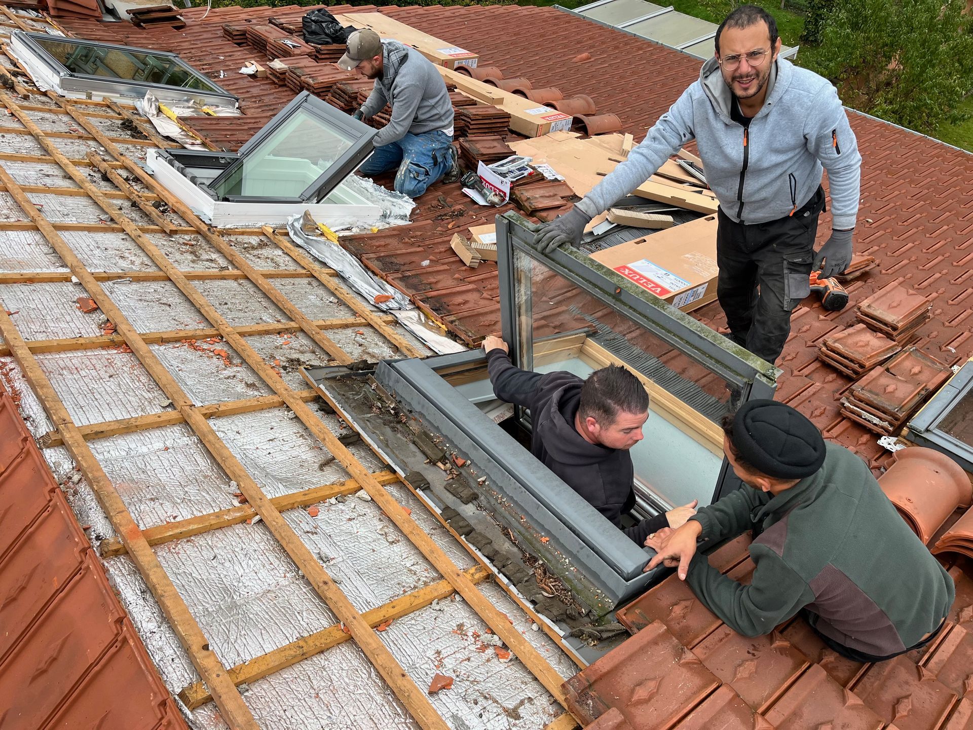 Des ouvriers du bâtiment installent des puits de lumière sur un toit de tuiles. Trois hommes travaillent activement : l’un désigne du doigt, les autres tiennent ou fixent le verre.
