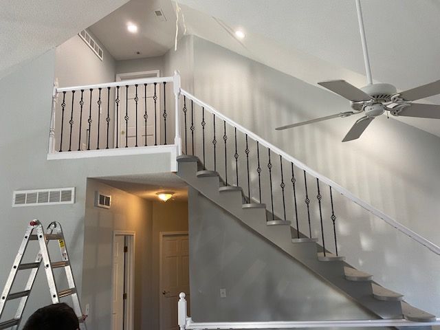 A person is standing on a ladder in a room with stairs and a ceiling fan.