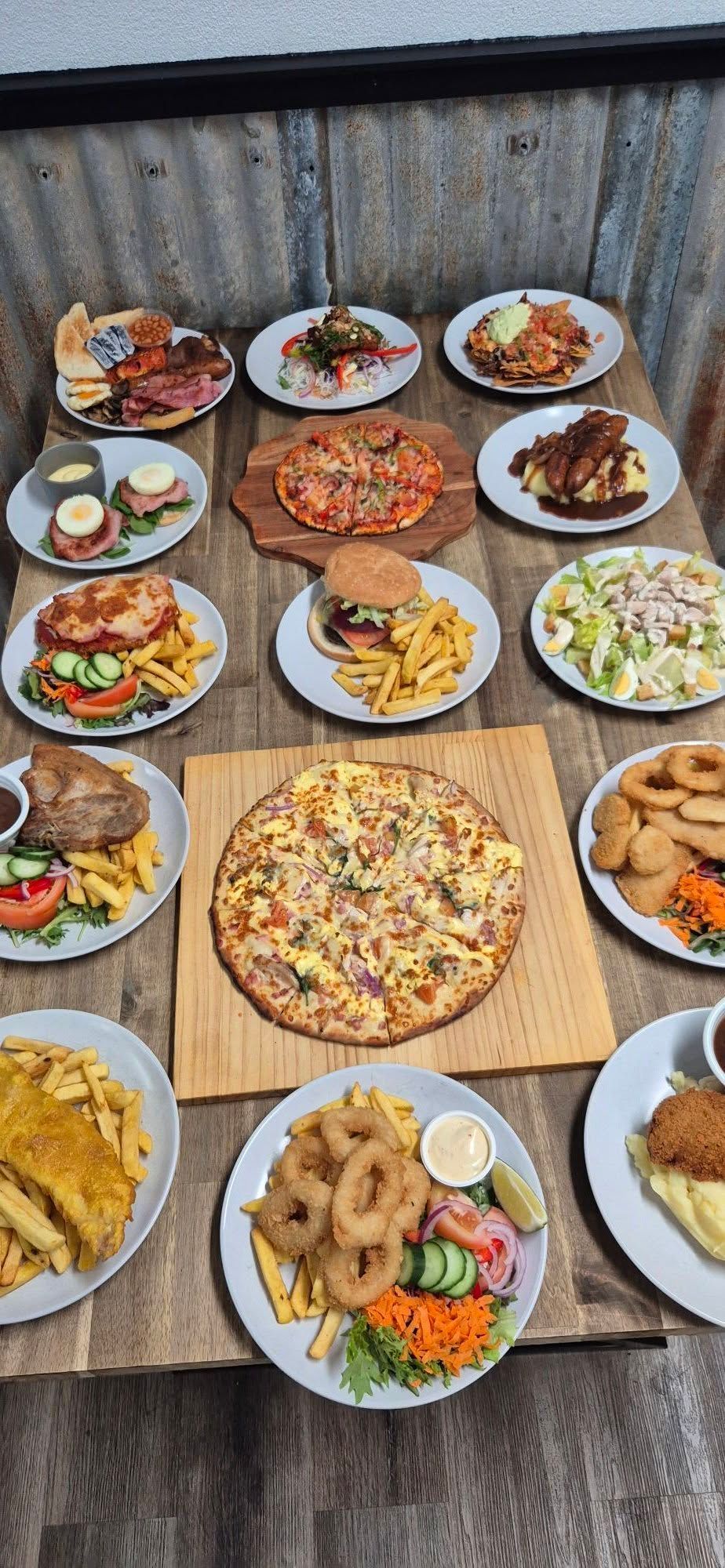Assortment of various dishes, including pizza, burgers, fish and chips, calamari rings, and salad. Set on a wooden table.
