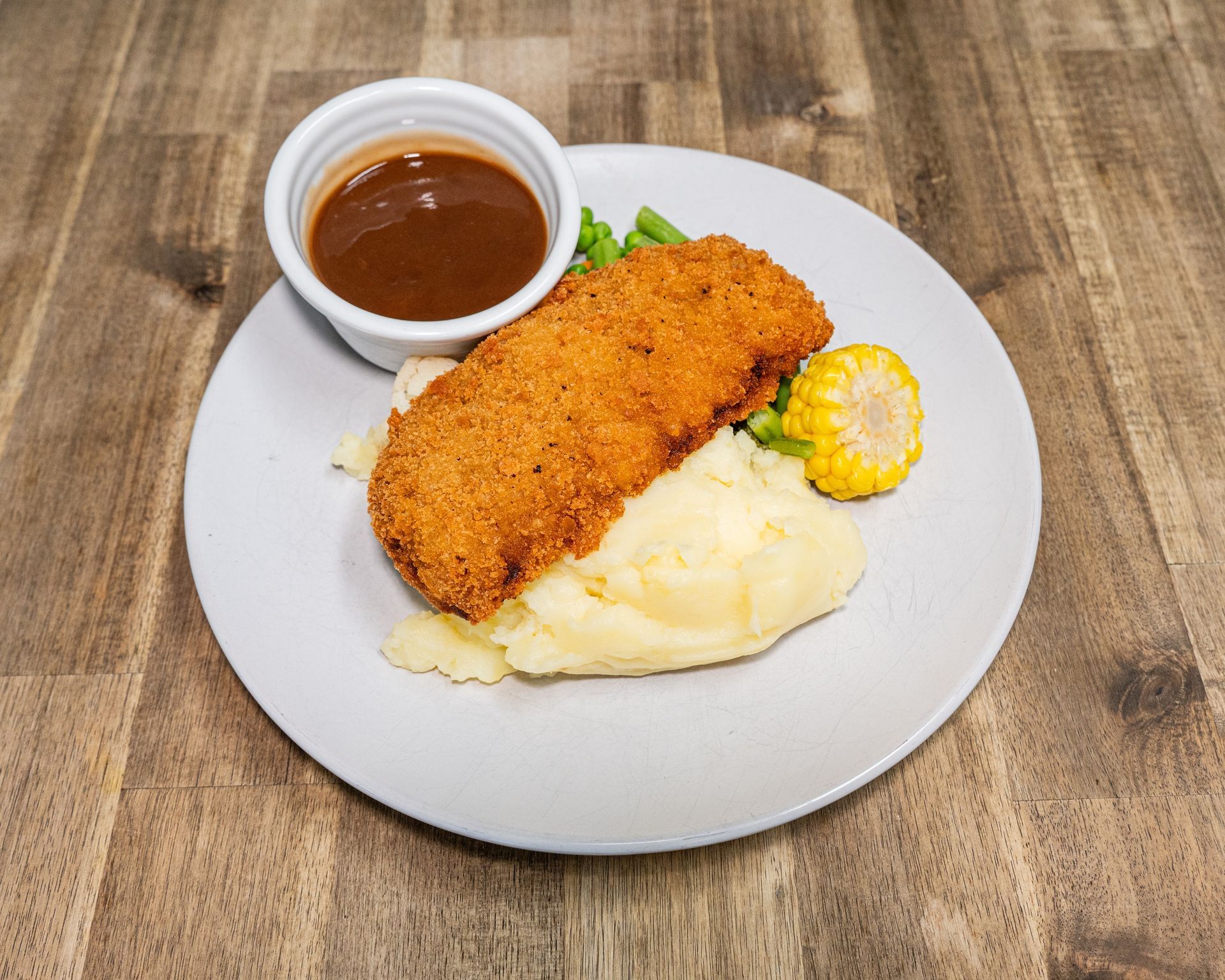Breaded cutlet with mashed potatoes, gravy, vegetables on a plate, wooden table.