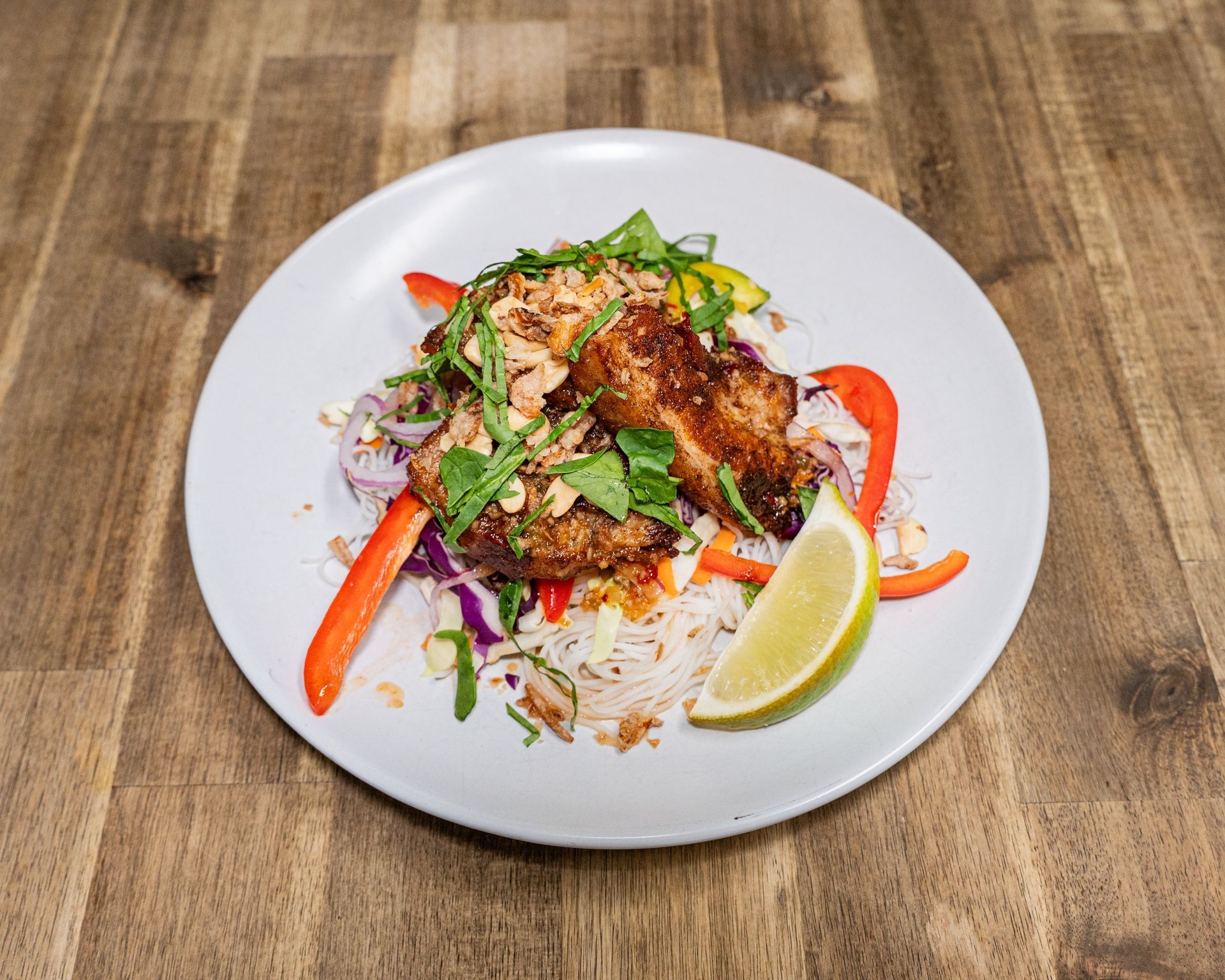 Chicken dish with vegetables, peanuts, lime wedge, on a white plate atop a wood table.