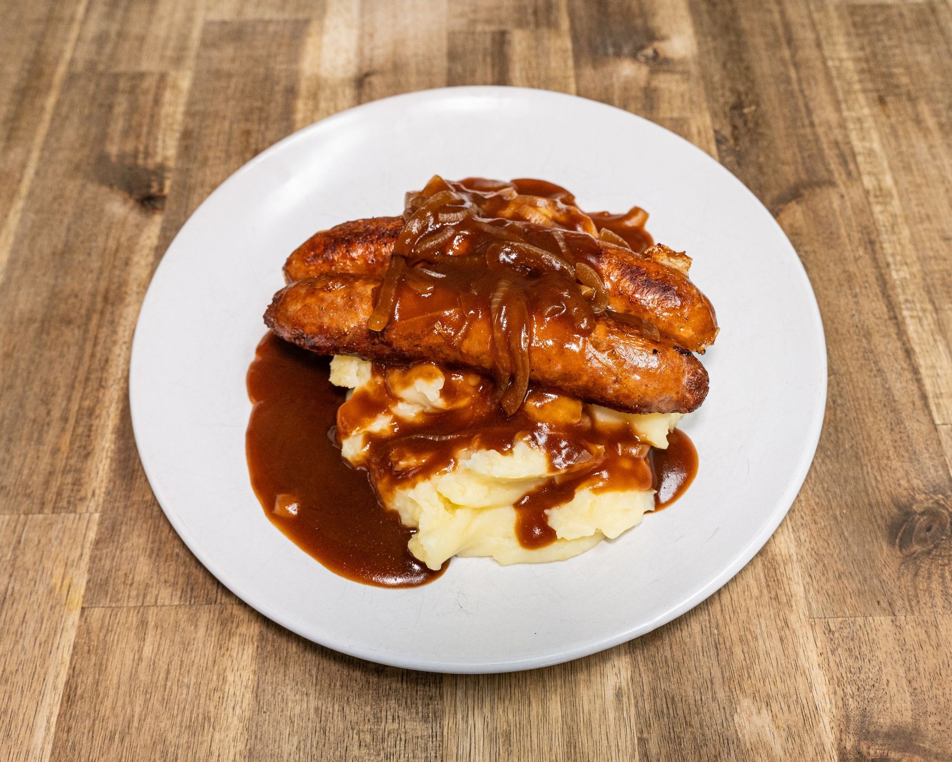 Sausages with mashed potatoes, covered in gravy, on a white plate atop a wooden table.