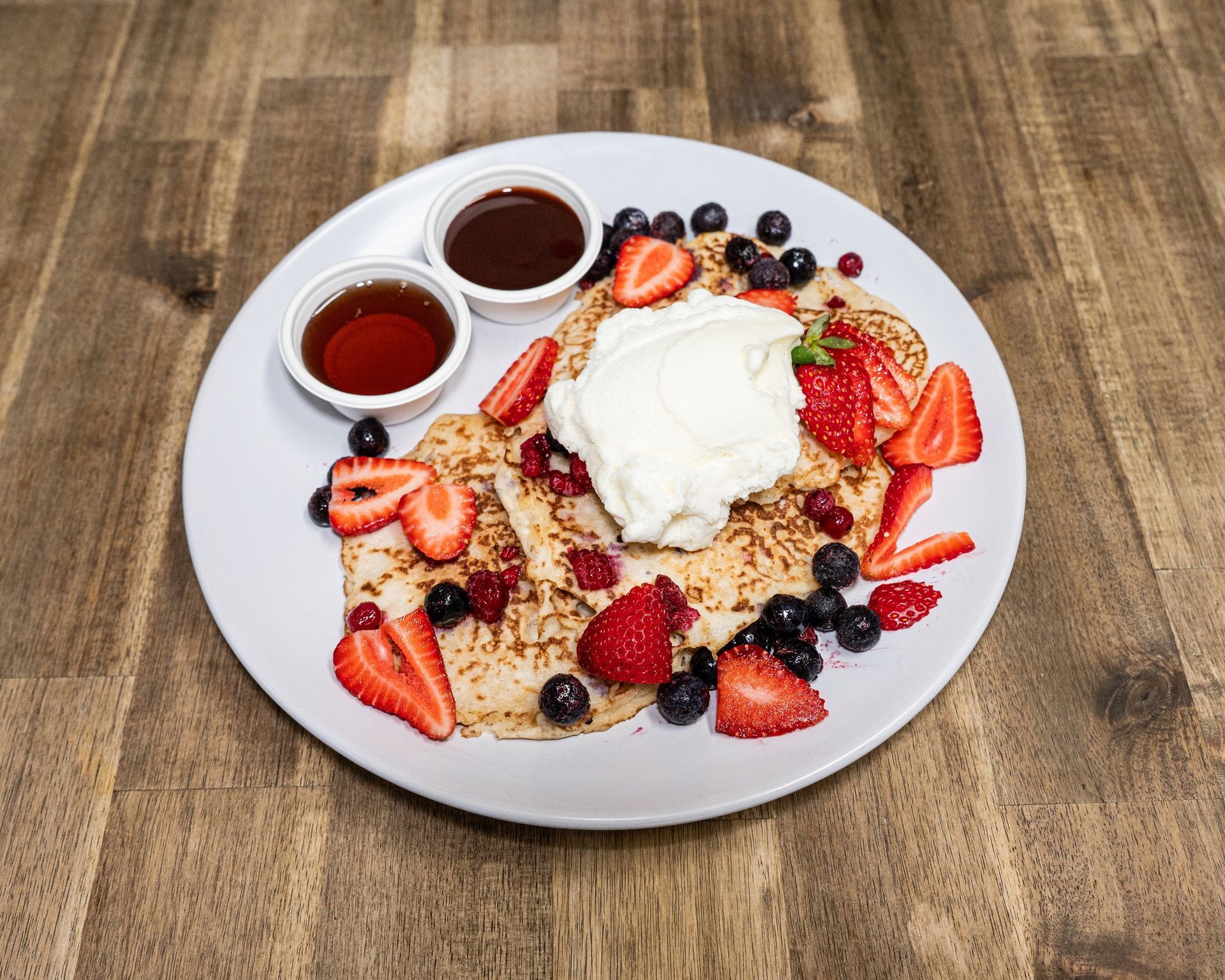 Pancakes topped with berries and whipped cream, served with syrup and chocolate sauce on a white plate.