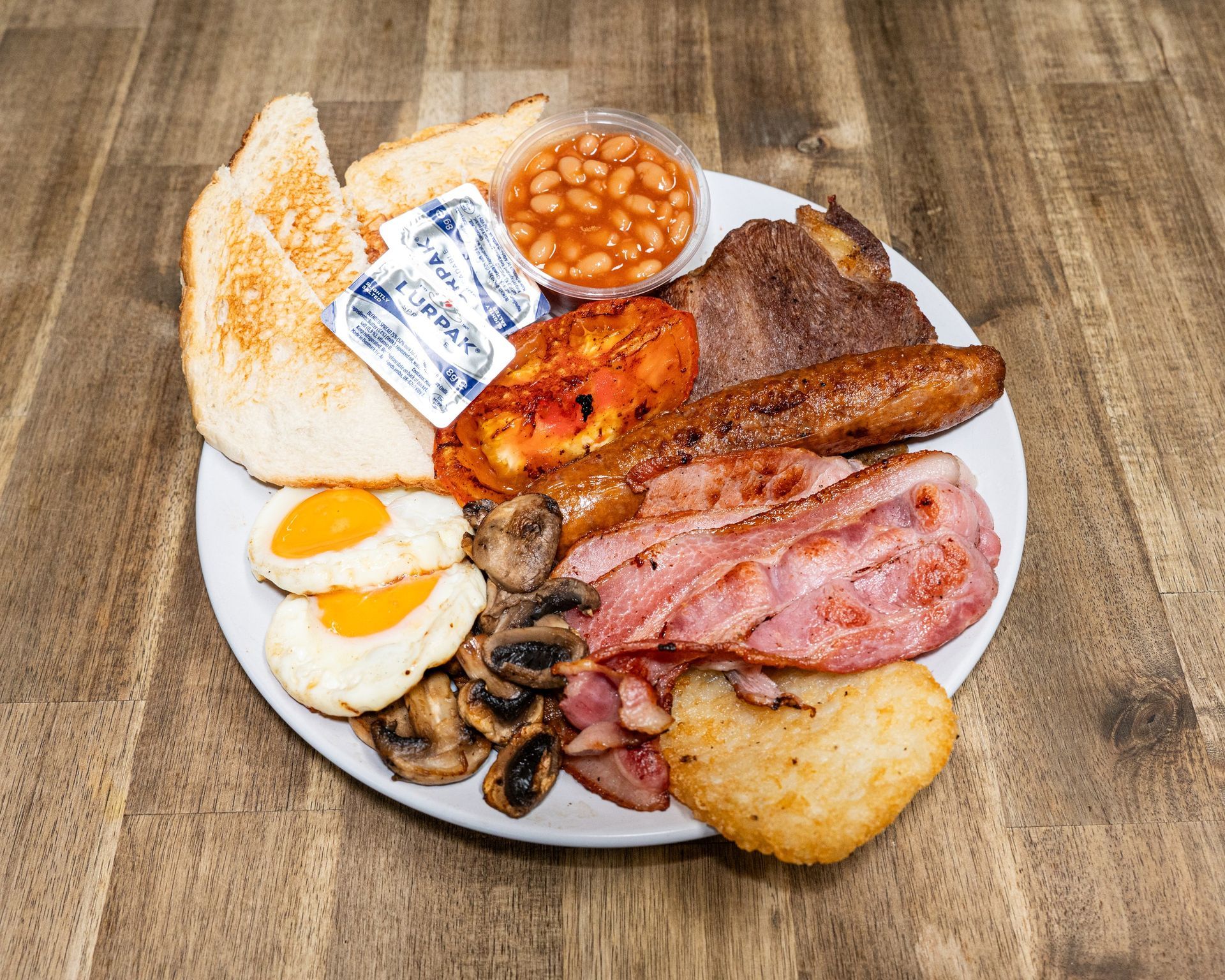 Full English breakfast on a white plate: fried eggs, bacon, sausage, beans, hash brown, toast, tomato, mushrooms.