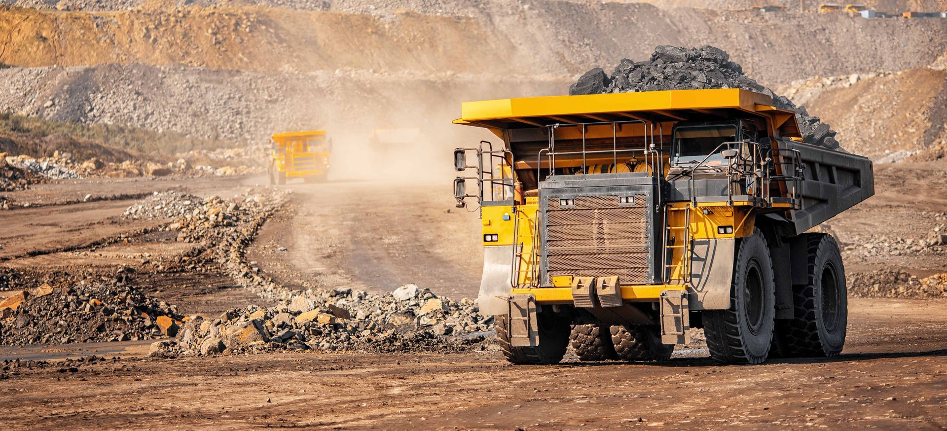 Yellow mining truck loaded with dark rocks, in a quarry. Another truck visible in the distance.