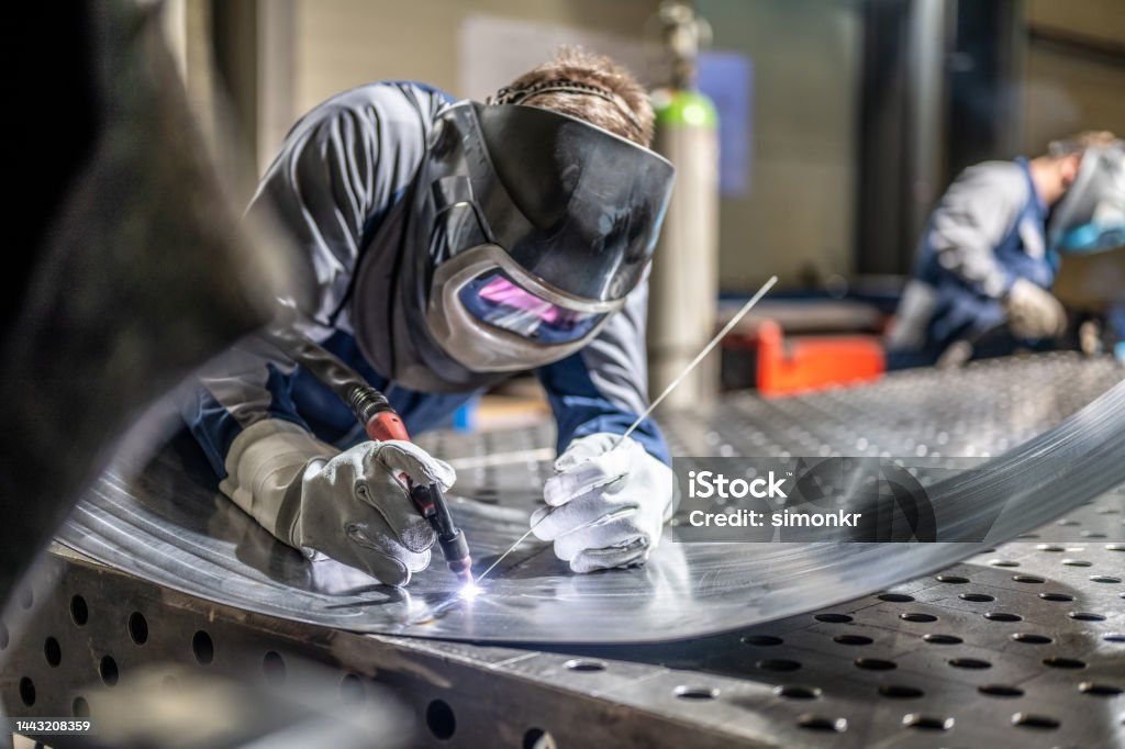 Welder in protective gear welding metal sheet on a workshop table. Another welder in the background.