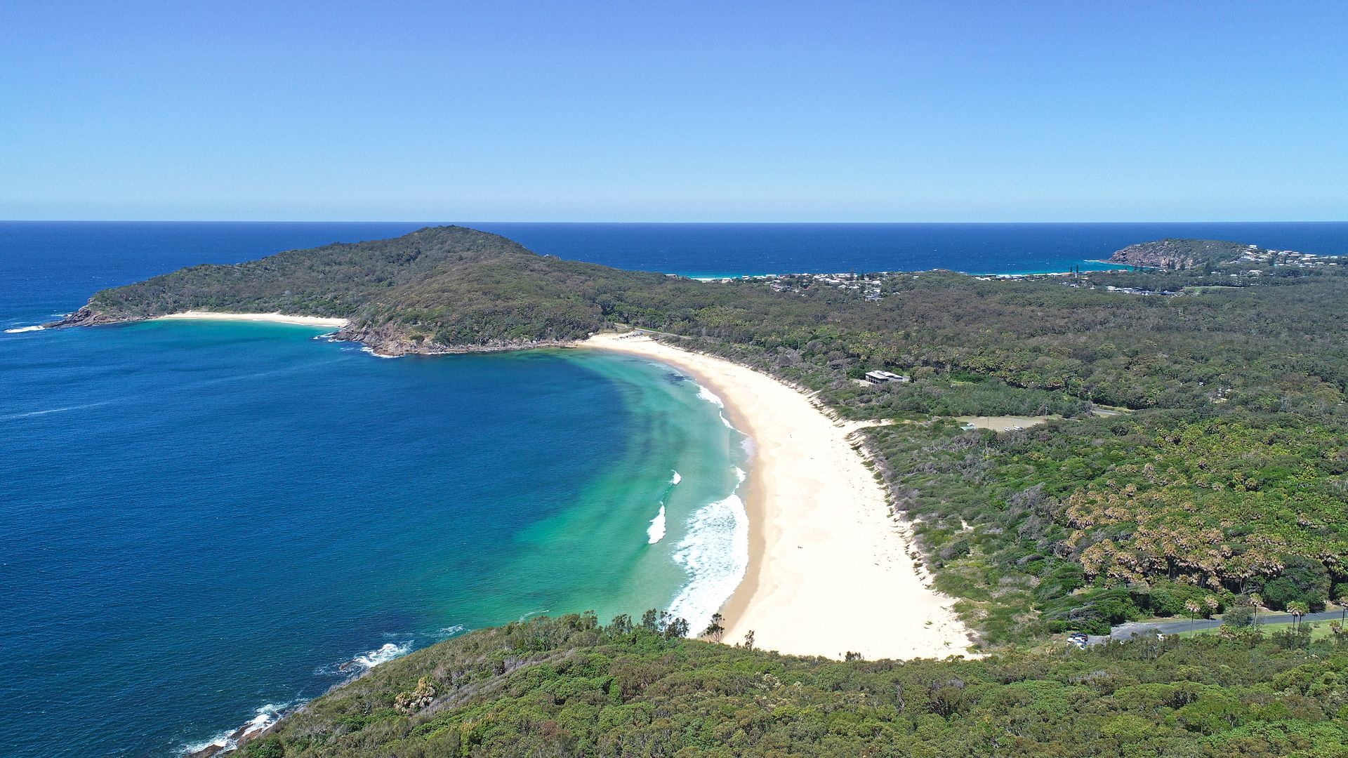 Aerial view of a sandy beach, turquoise water, and lush green forest under a clear blue sky.— Pacific Palms Caravan Park in Elizabeth Beach, NSW
