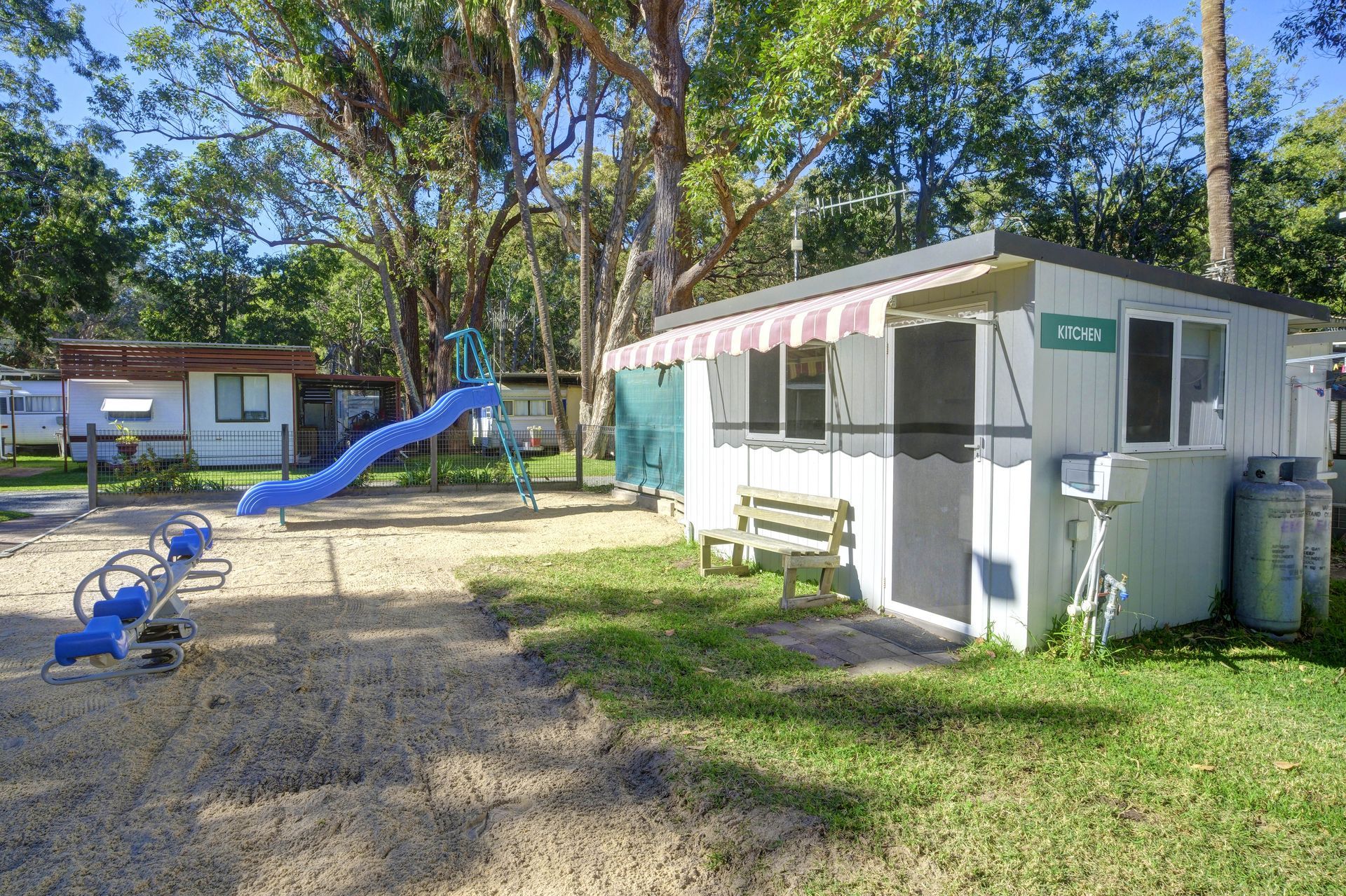 Playground with slide, seesaw, and small white building with striped awning. Sand and grass are visible.— Pacific Palms Caravan Park in Elizabeth Beach, NSW