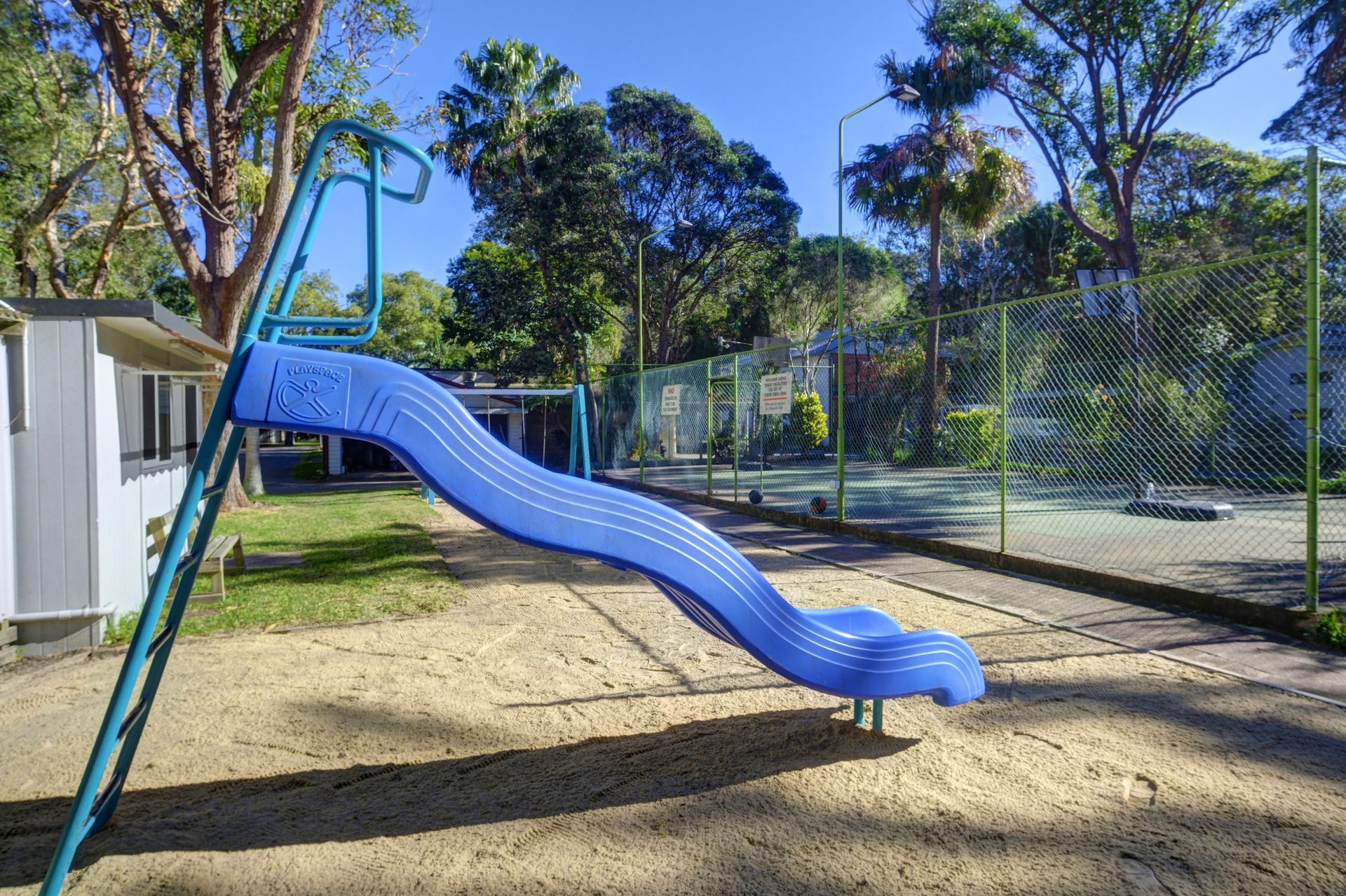 Blue playground slide on a sand surface, green grass, trees, and fence in the background. Sunny day.— Pacific Palms Caravan Park in Elizabeth Beach, NSW