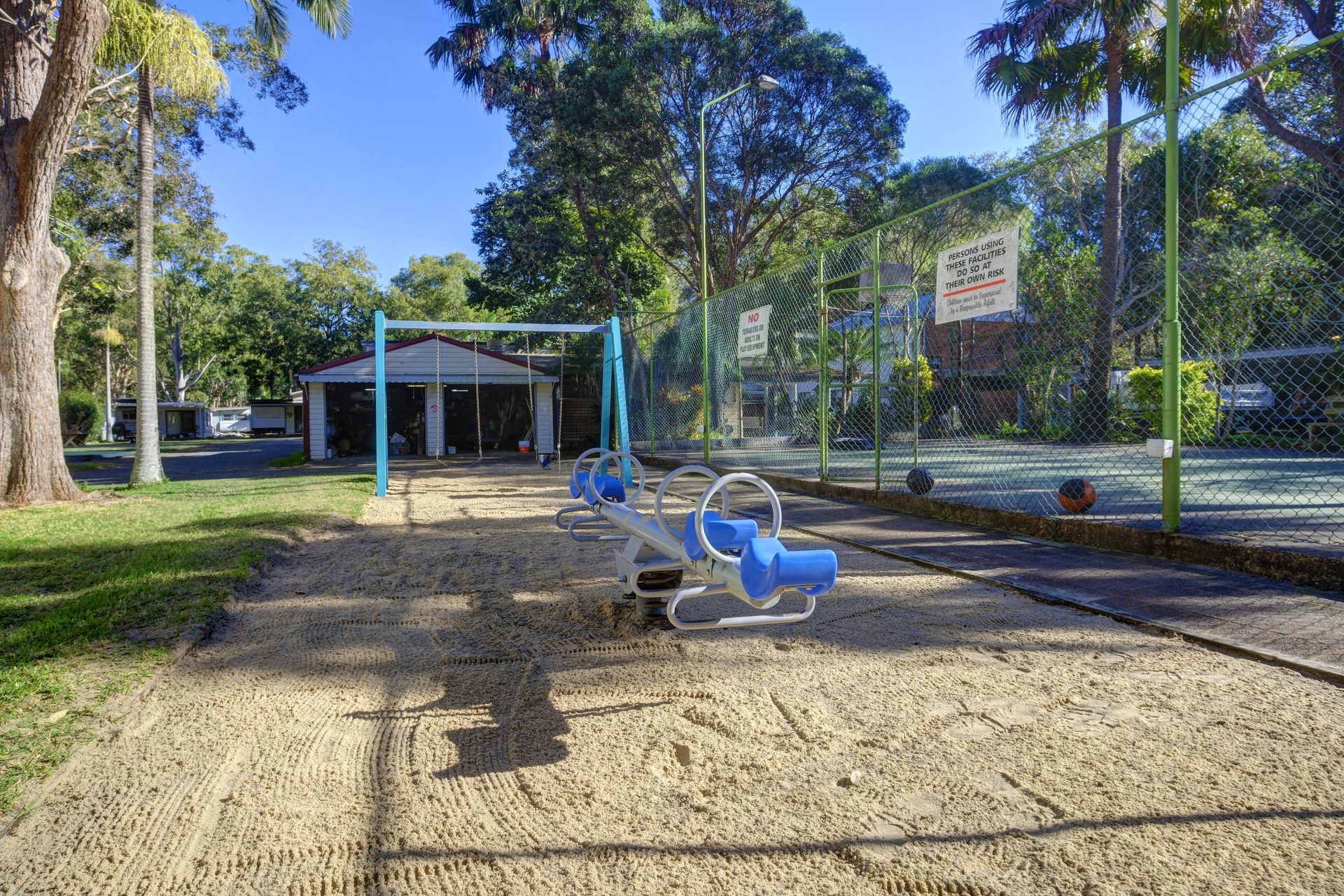 Playground with swings and a seesaw on a sand surface, fence, trees, and a shelter in the background.— Pacific Palms Caravan Park in Elizabeth Beach, NSW