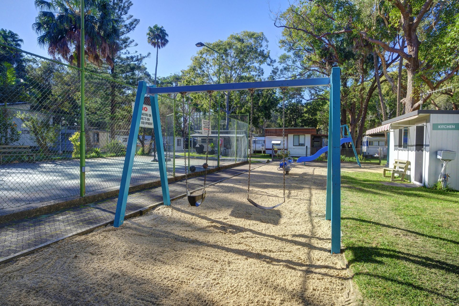 Playground with blue swing set on woodchip surface, next to a grassy area and fence.— Pacific Palms Caravan Park in Elizabeth Beach, NSW