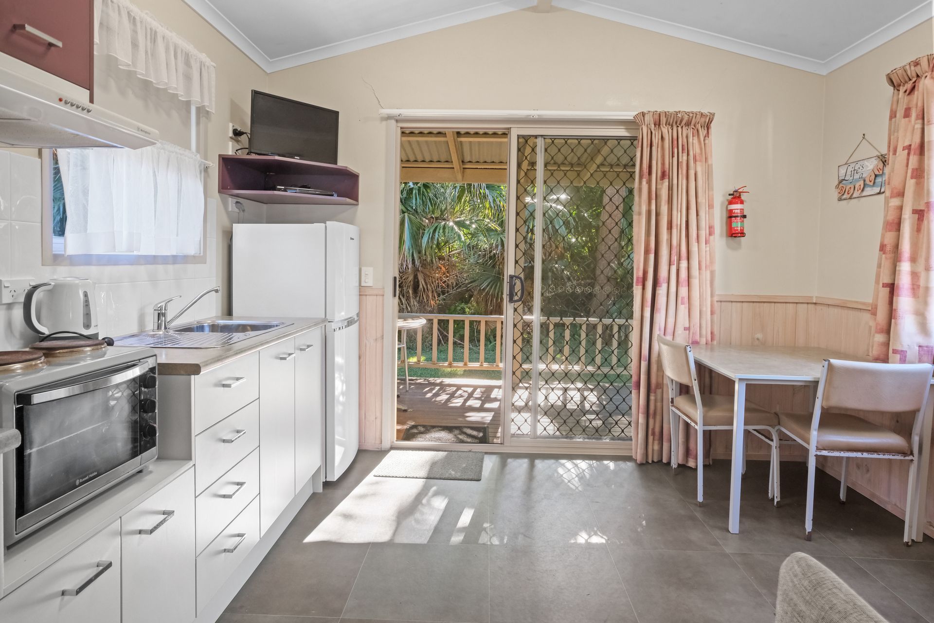 Small Kitchen and Dining Area With Sliding Door to Outside. White Cabinets, Refrigerator, Table, and Chairs — Pacific Palms Caravan Park in Elizabeth Beach, NSW