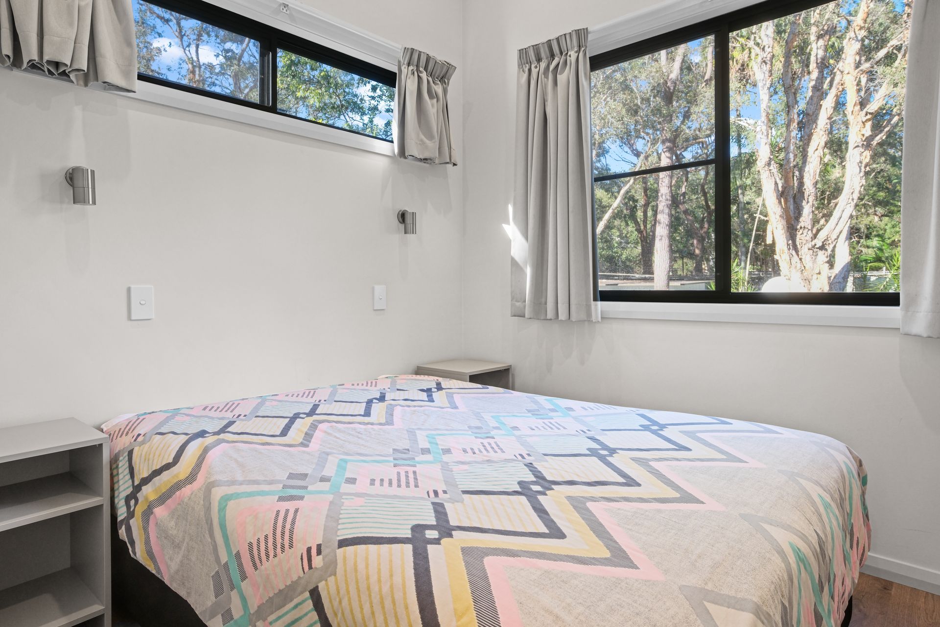 Bedroom with bed, windows with curtains, and bedside tables; sunlight enters.— Pacific Palms Caravan Park in Elizabeth Beach, NSW