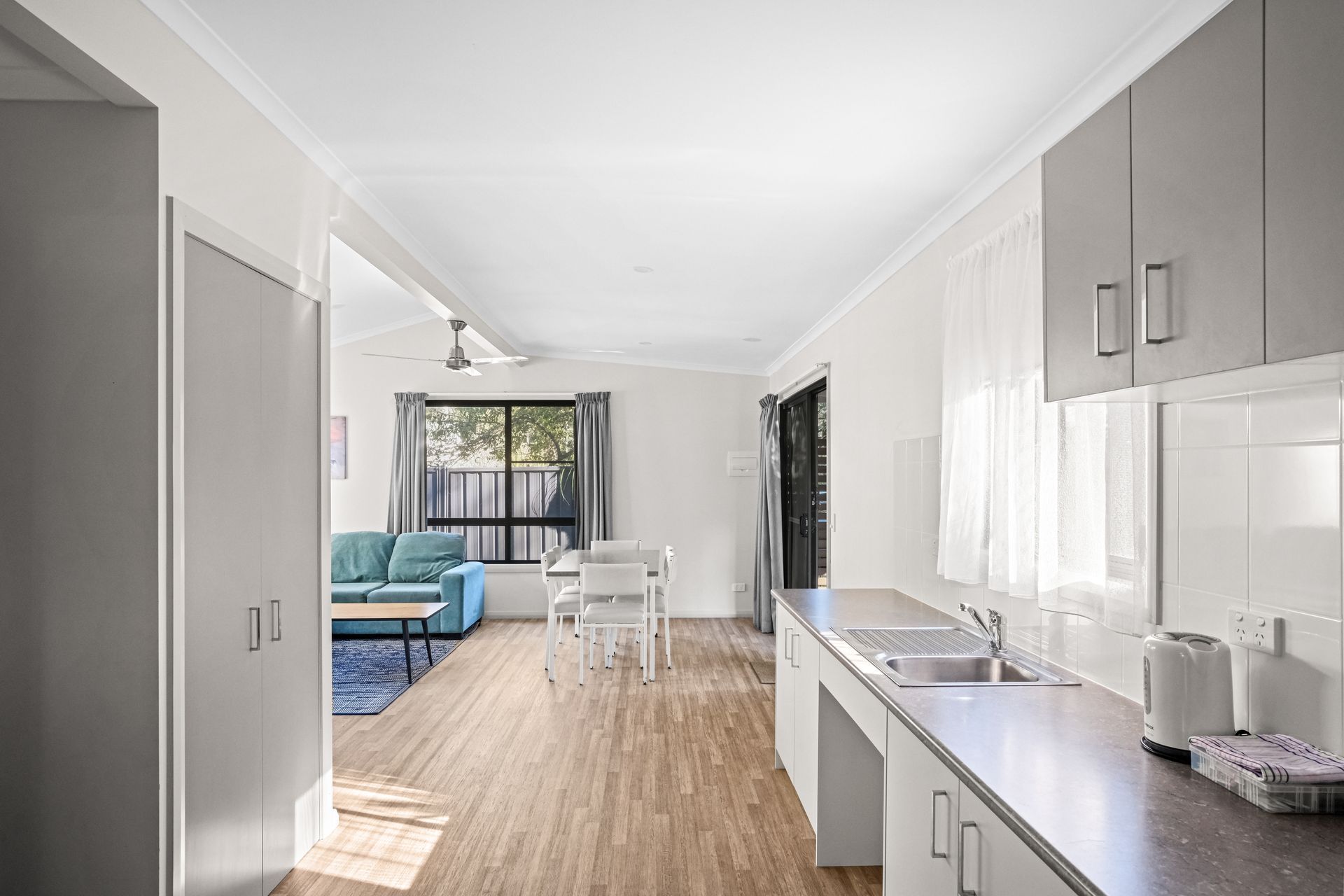 Kitchen with gray cabinets and countertops, leading to a living area with blue sofa and dining set.— Pacific Palms Caravan Park in Elizabeth Beach, NSW