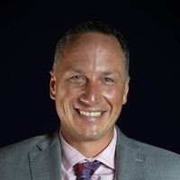 Man in a suit, smiling broadly, against a dark background. He is wearing a pink shirt and patterned tie.