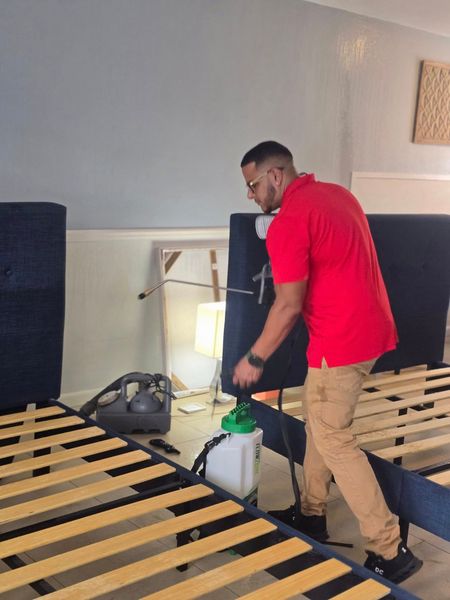 A person in a red shirt uses a handheld sprayer to treat dark upholstered headboards in a room with bed frames.
