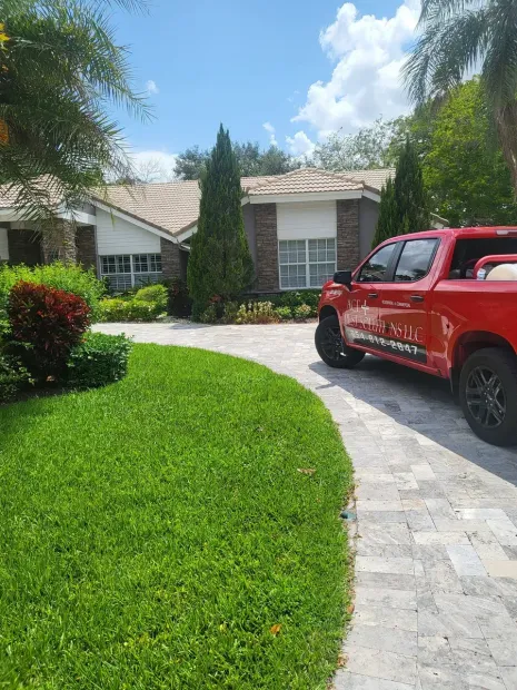A red pickup truck is parked on a paved driveway in front of a house with a tile roof, lawn, and surrounding trees.