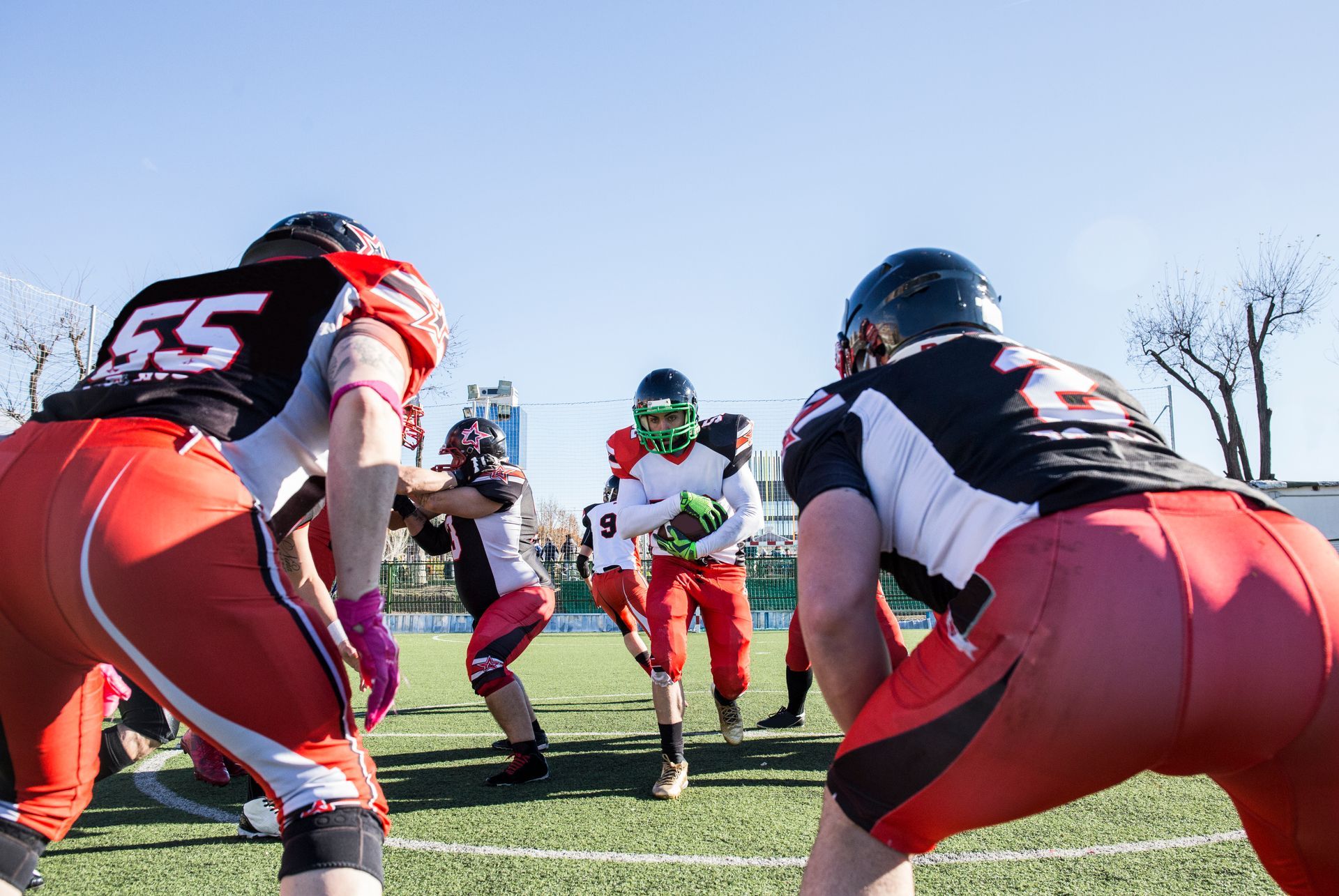 Football players in red and black uniforms on a green field. A player runs with the ball, others are in a scrimmage.