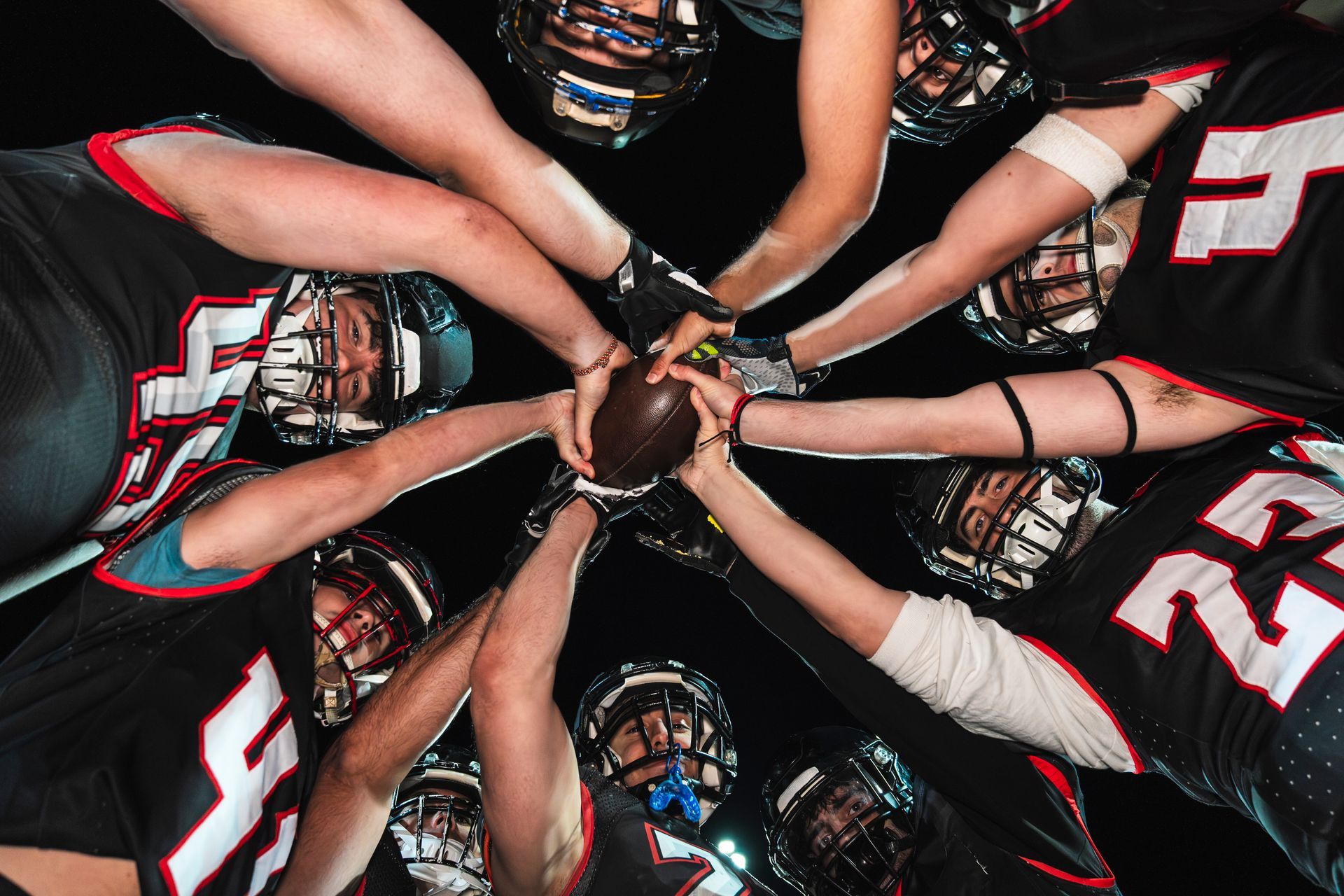 Football players huddle around a football, hands joined, wearing black uniforms and helmets.