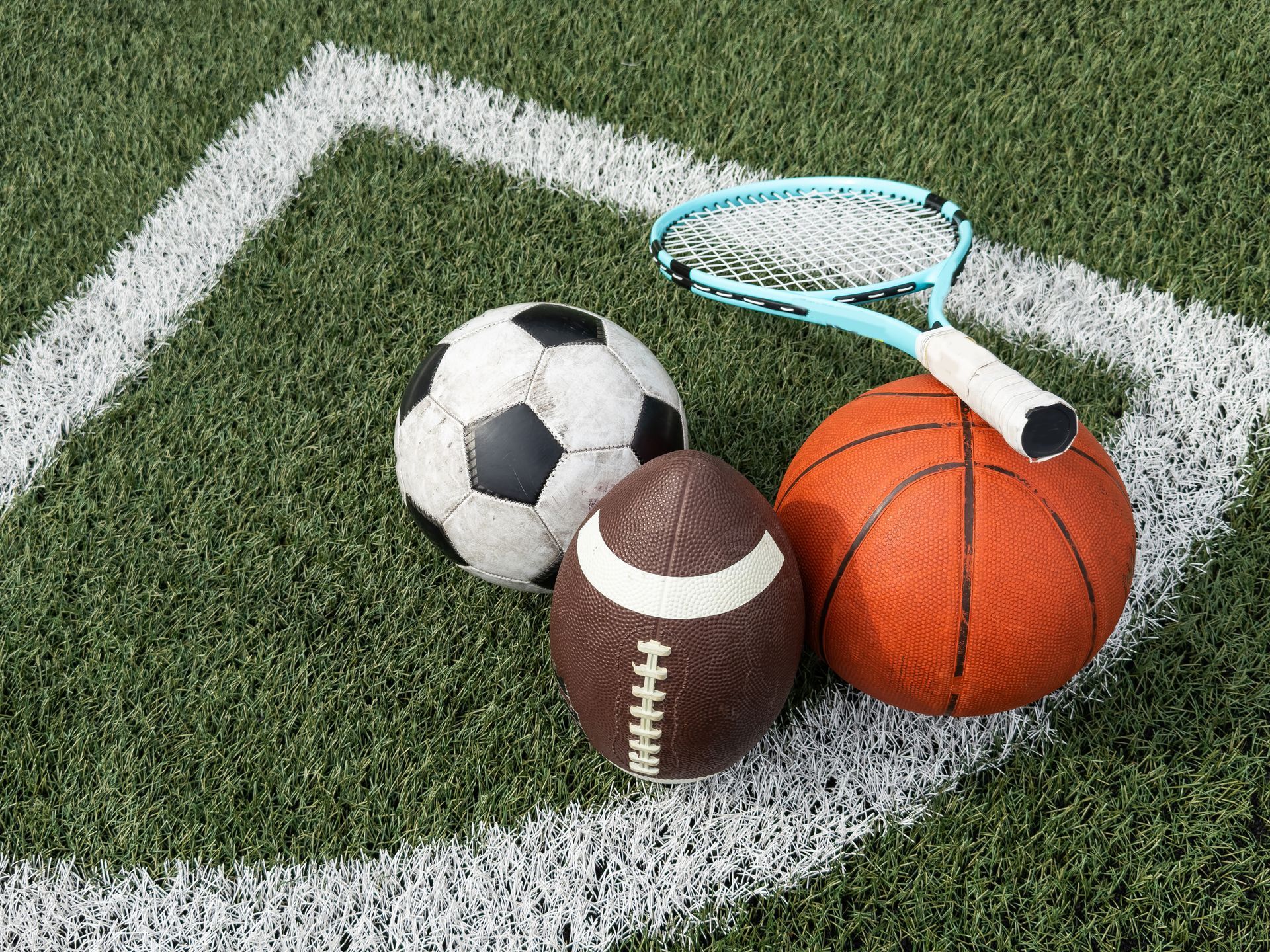 Sports equipment: soccer ball, football, basketball, and tennis racket on a field.