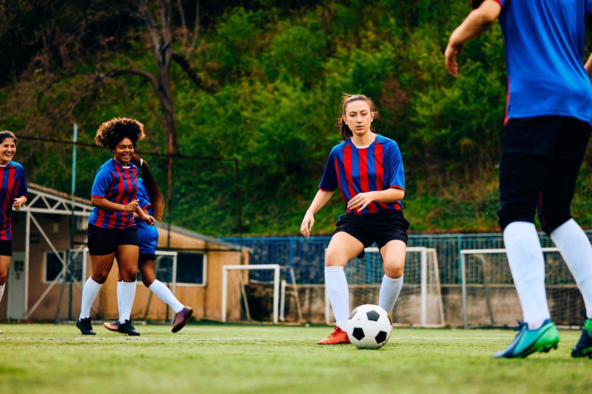 Soccer players on field, one dribbles ball. Others in blue and red jerseys near goals and trees.