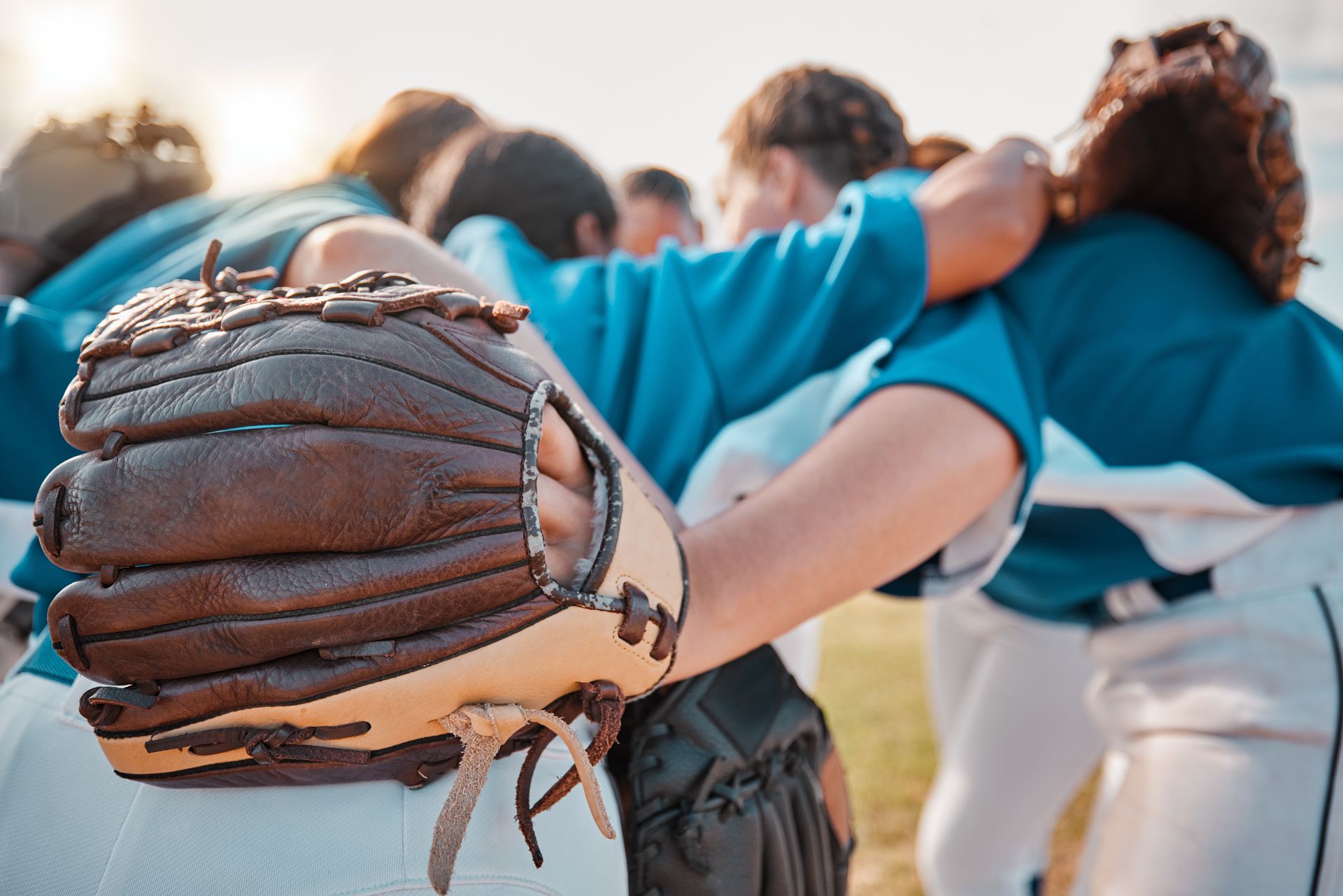 Baseball glove in focus, team huddle in the background, blue shirts, outdoor setting.