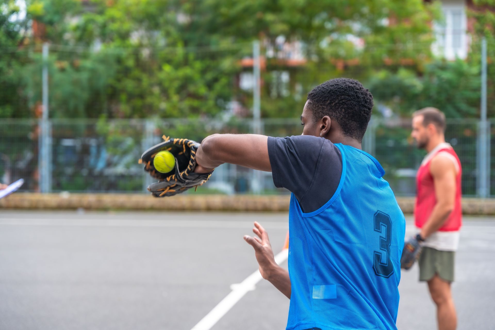 A person in blue jersey throws a softball on an outdoor court, another person in the background.