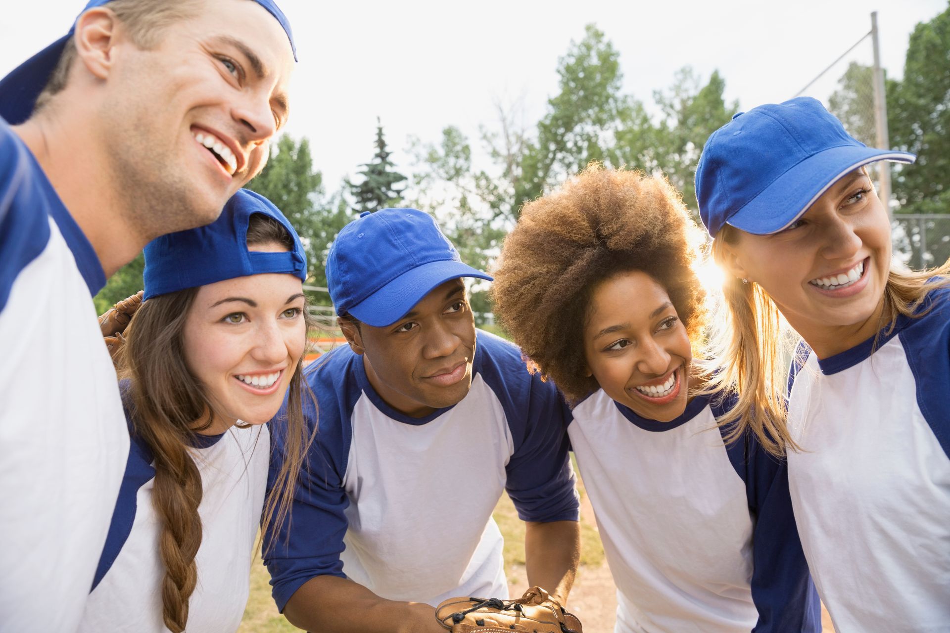 Five people in baseball uniforms smile outdoors, holding a small cake.