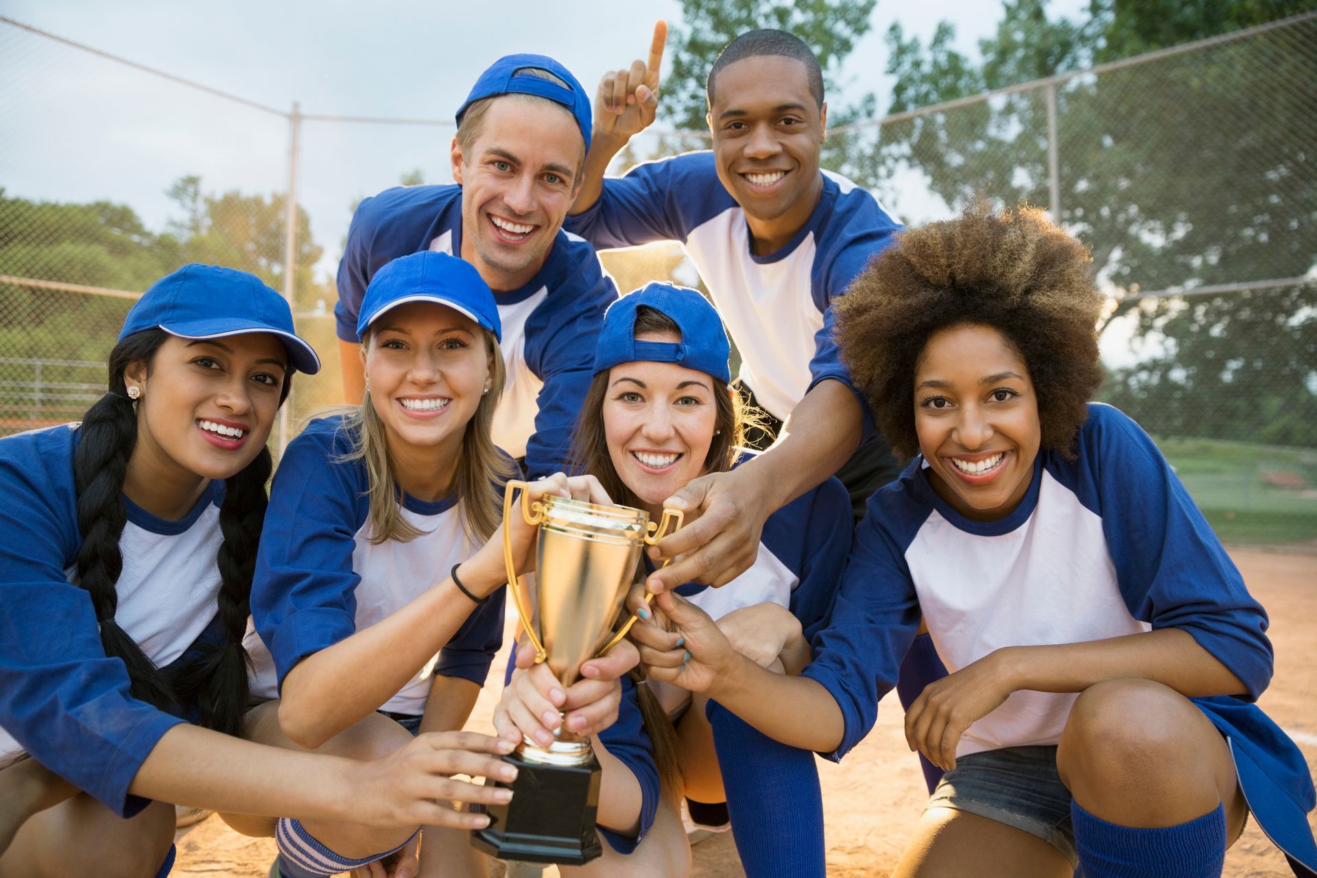 Softball team in blue and white uniforms celebrates, holding a trophy and smiling on a field.