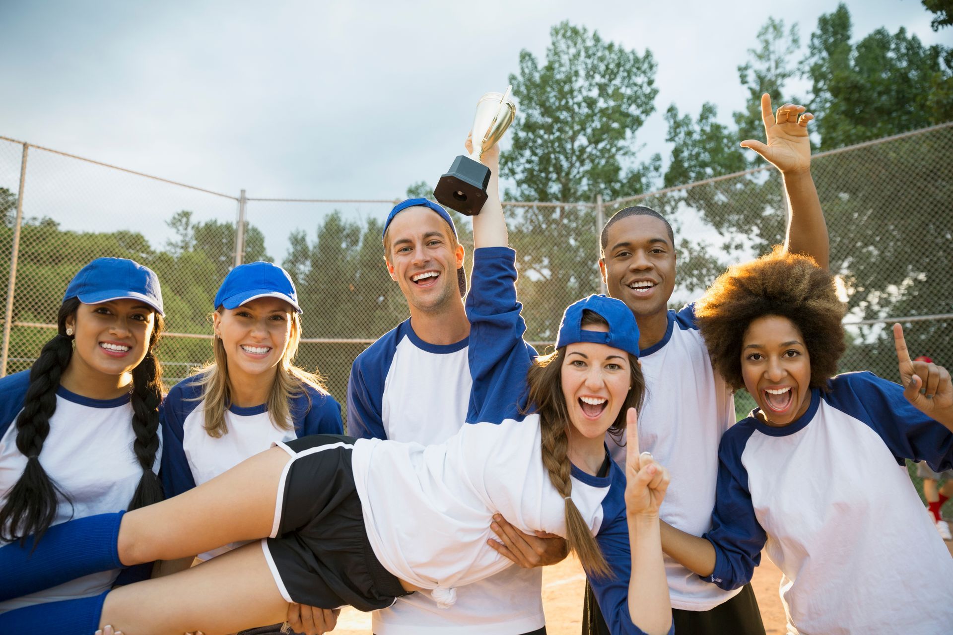 Softball team celebrates victory, holding trophy and lifting a teammate.