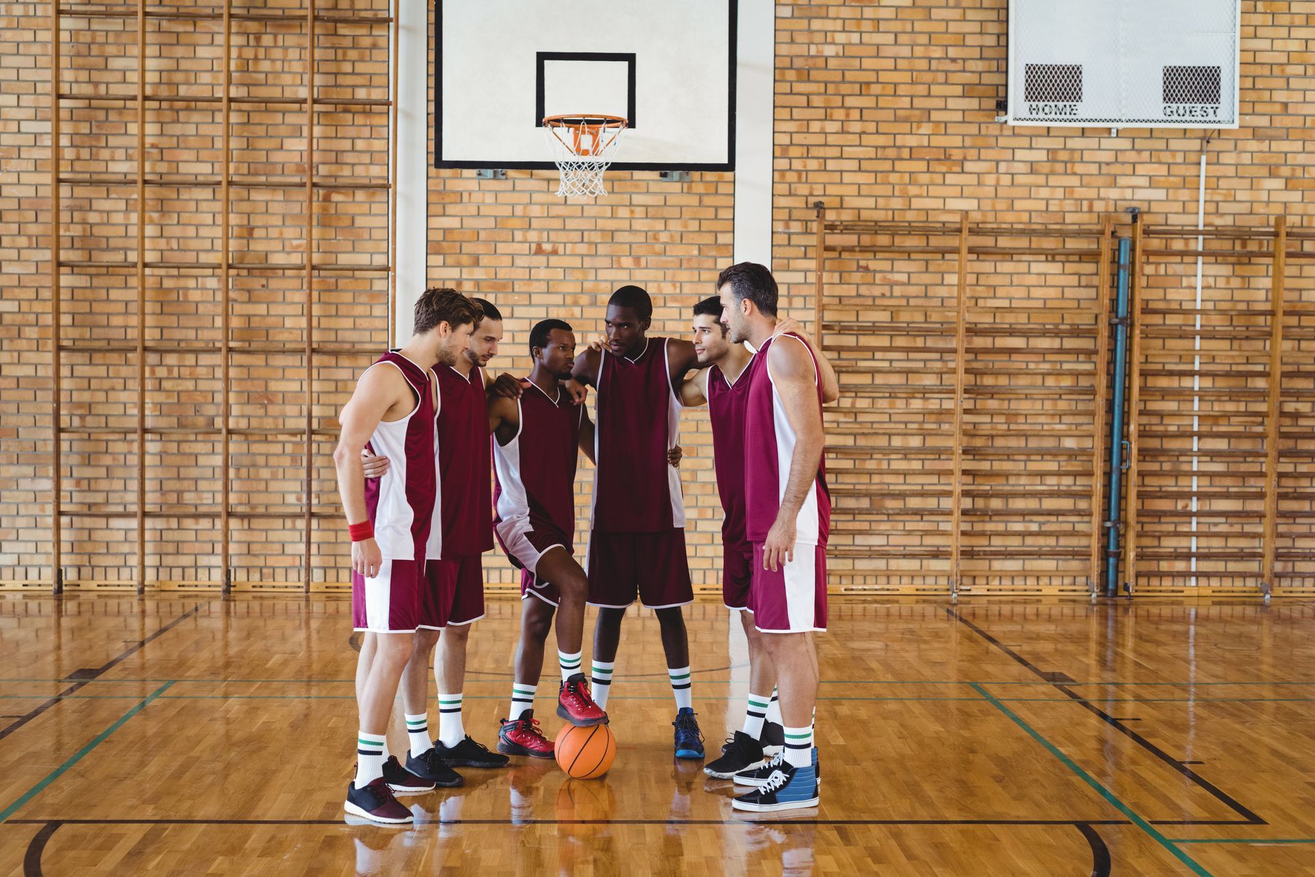 Basketball team in maroon uniforms huddles on a wooden court, ball at their feet, near a hoop.