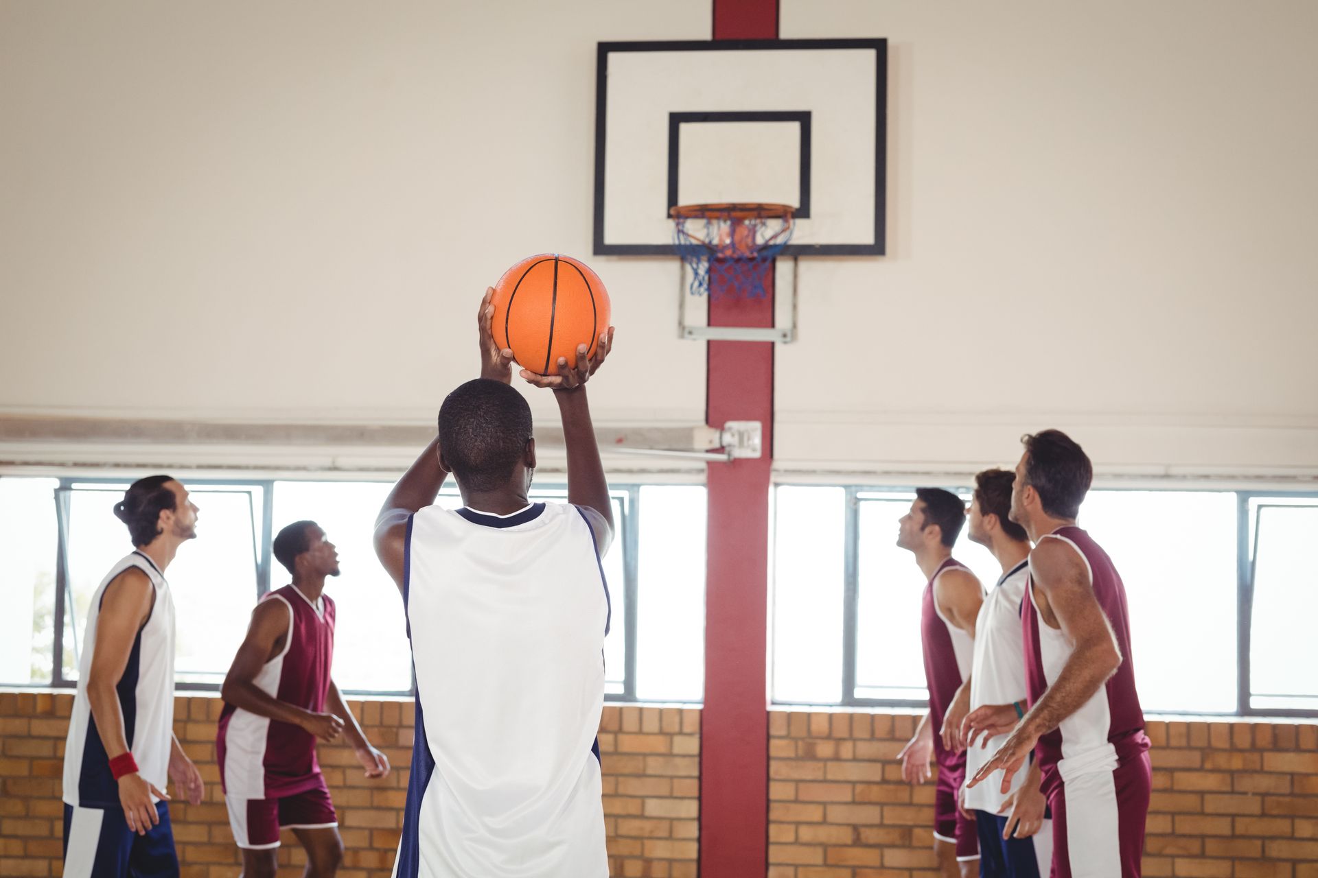 Basketball player shooting, teammates watching. Court with hoop, red brick wall, and windows.