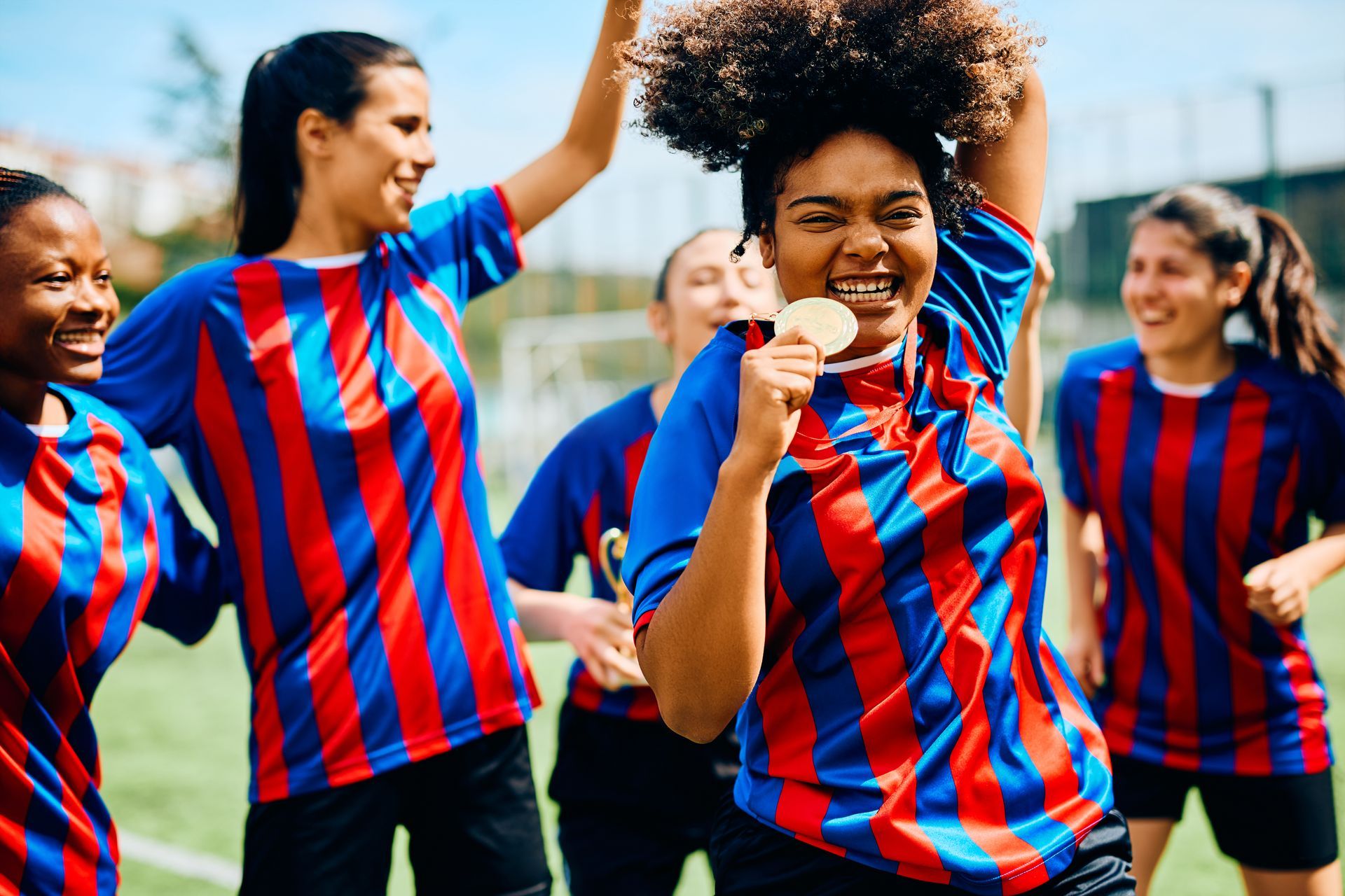 Soccer team celebrates victory, holding up a medal. Players wear blue and red striped jerseys, smiling on a field.